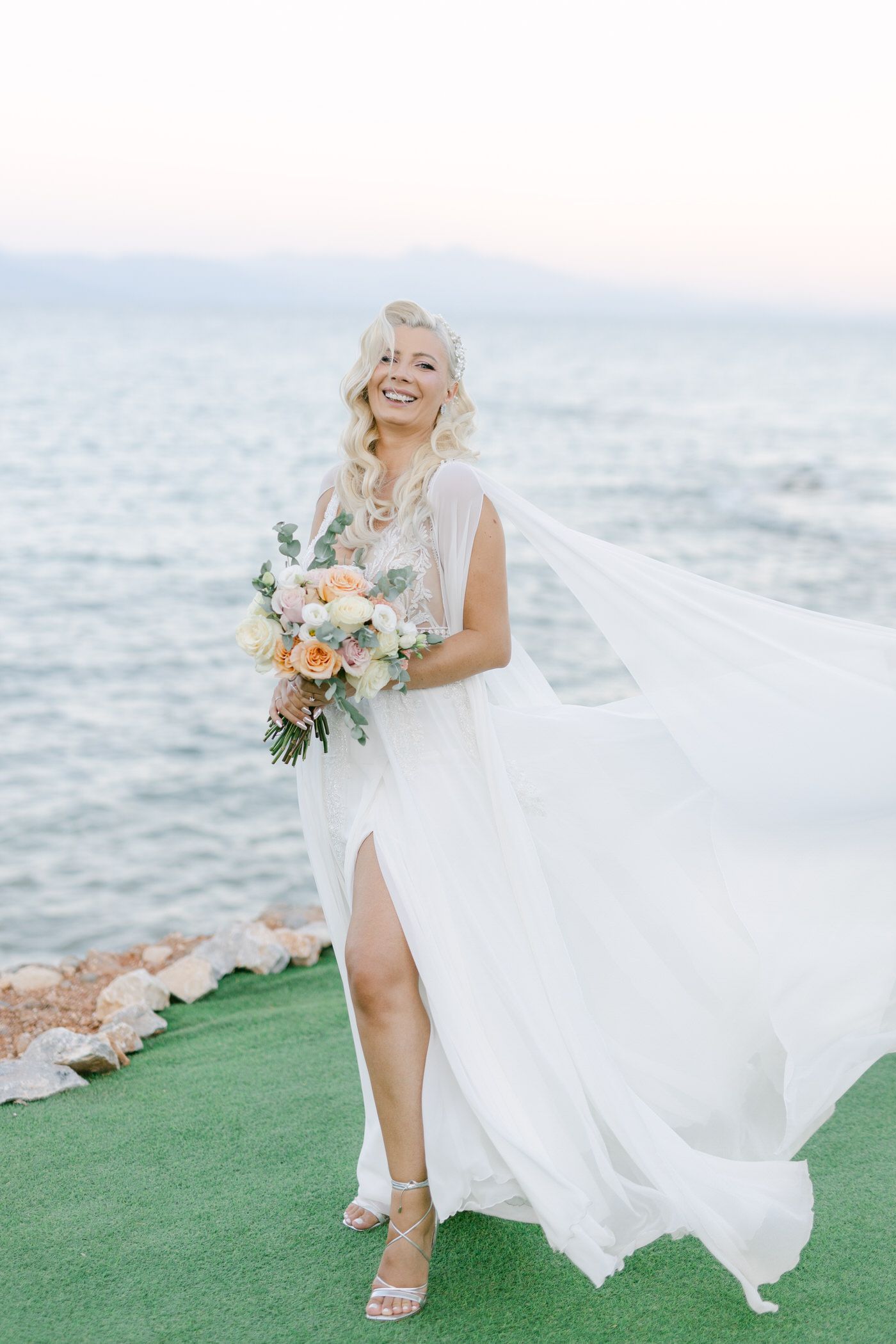 A bride in a flowing white dress with slit holds a peach and white bouquet while standing by the ocean.