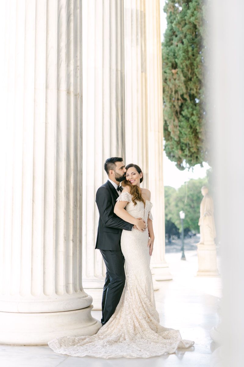 A couple shares a romantic moment among elegant white columns in a classical architectural setting with trailing greenery.