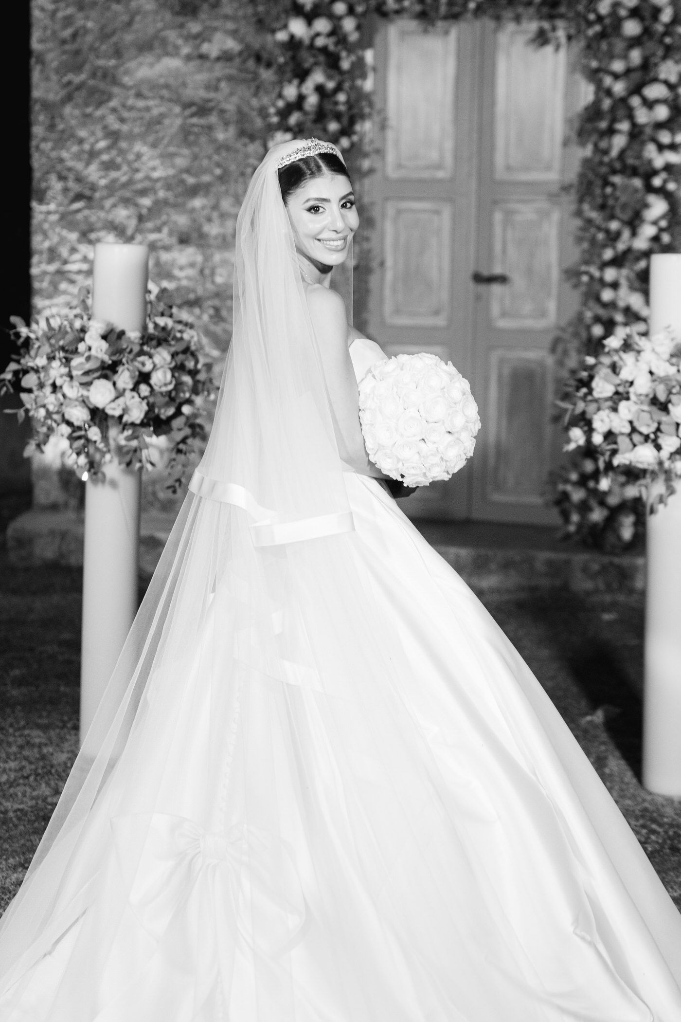 A bride in a classic white ballgown and long veil poses with her white bouquet against a floral-decorated doorway.