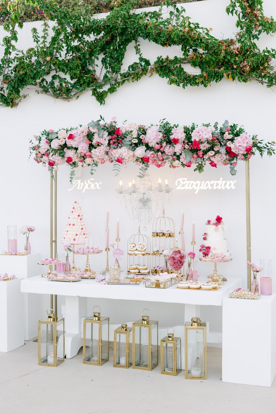 Luxurious dessert table with pink floral arrangements and gold lanterns beneath climbing vines.