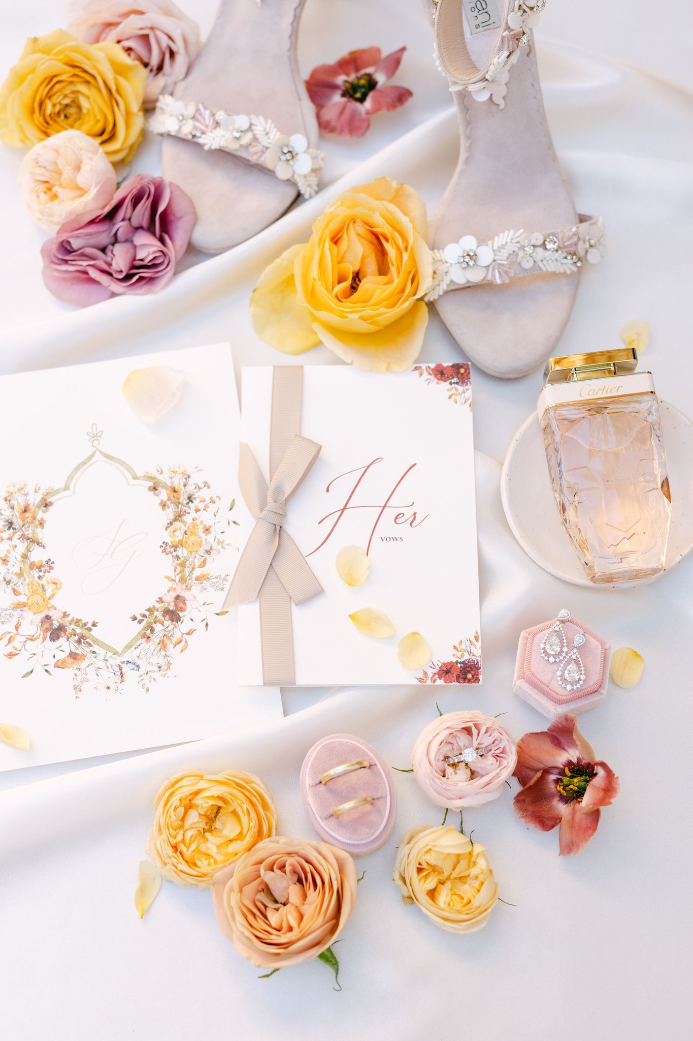 Wedding details flatlay with yellow and pink roses, elegant shoes with floral accents, and invitation with silk ribbon.