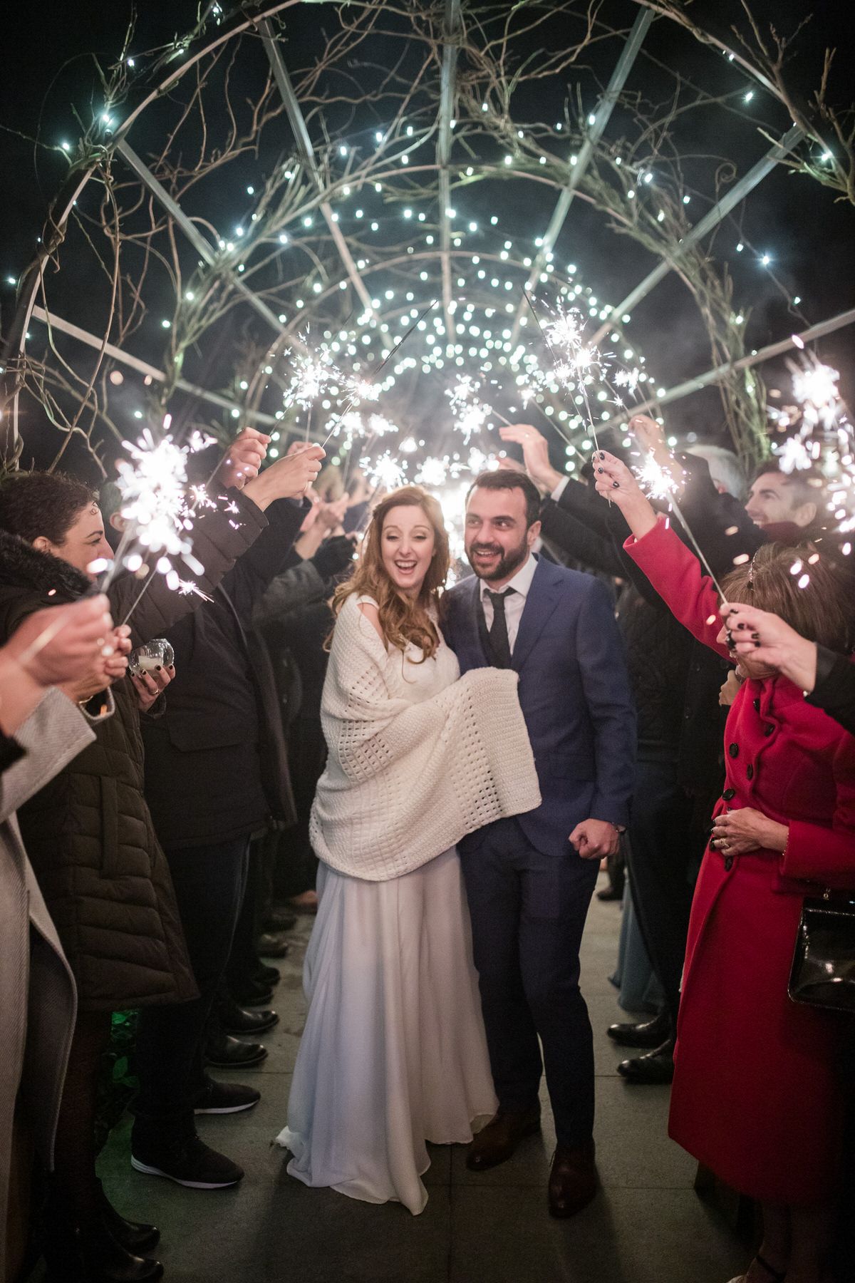 Wedding couple walks through sparkler send-off tunnel with guests holding sparklers under twinkling lights at night.