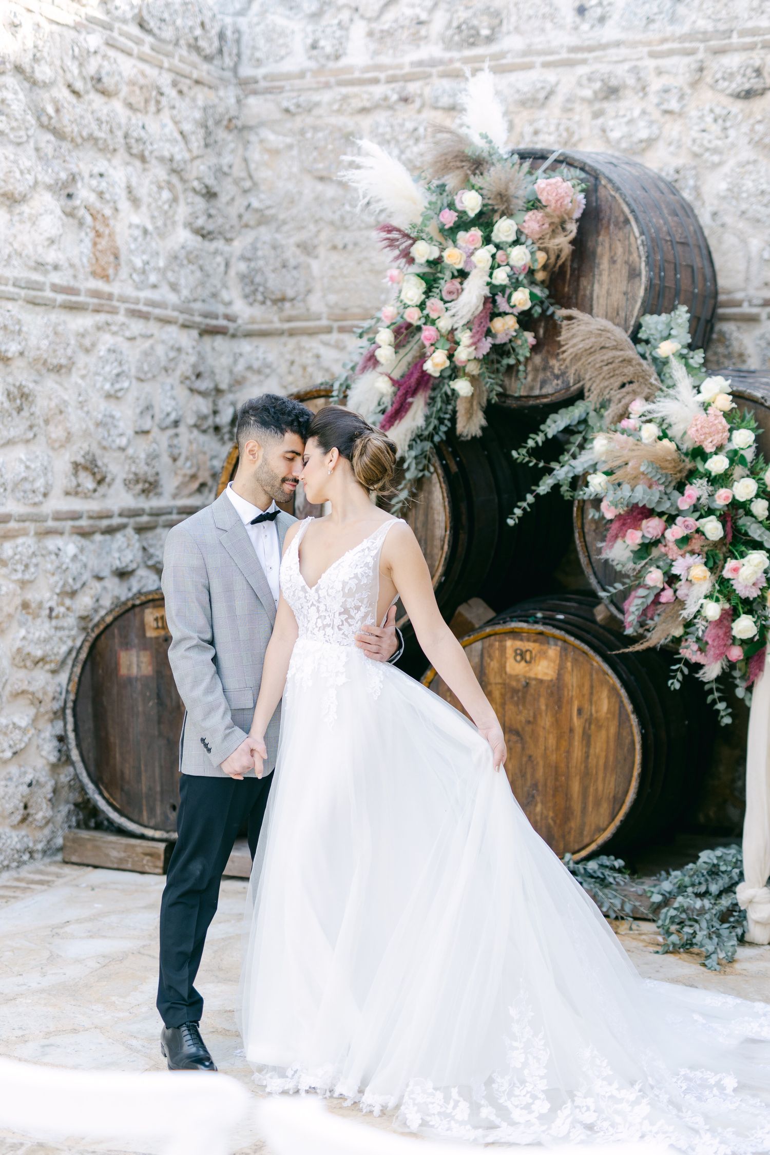 Newlyweds share an intimate moment against rustic wine barrels decorated with floral arrangements.
