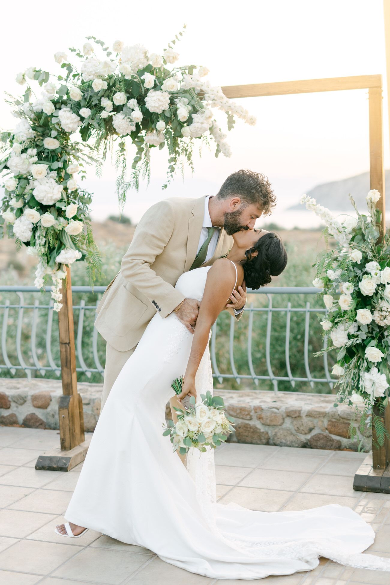 A romantic dip pose under an elegant white floral wedding arch at sunset.