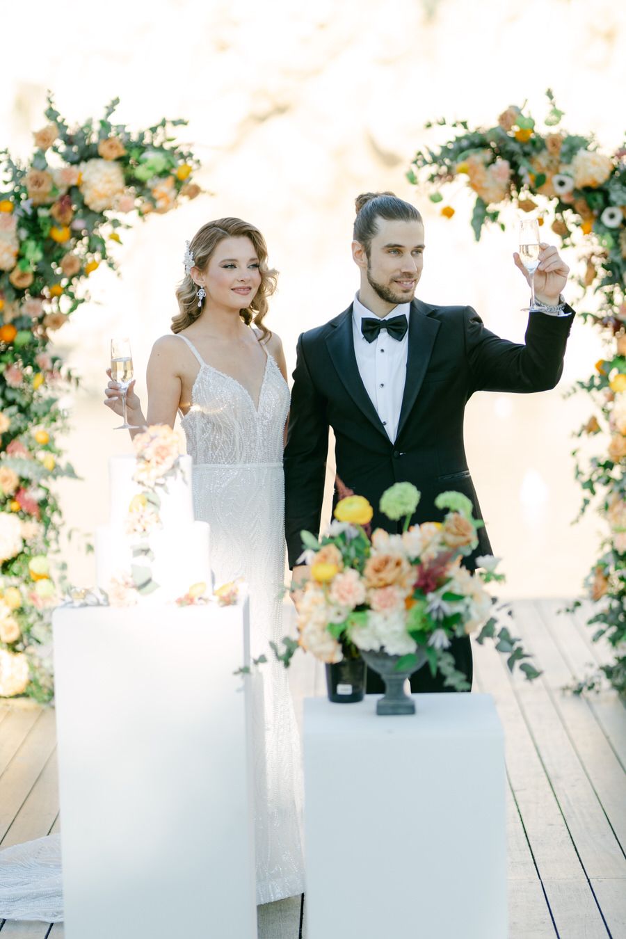Couple in wedding attire raising champagne glasses for a toast under floral arch decorations.