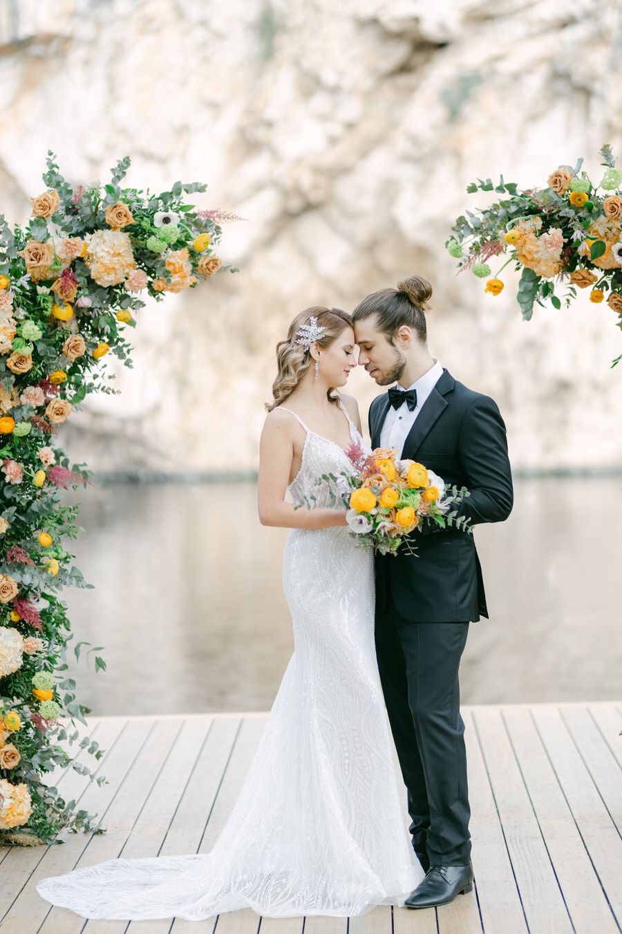 A couple shares an intimate moment under floral arches on a wooden dock by cliffs.