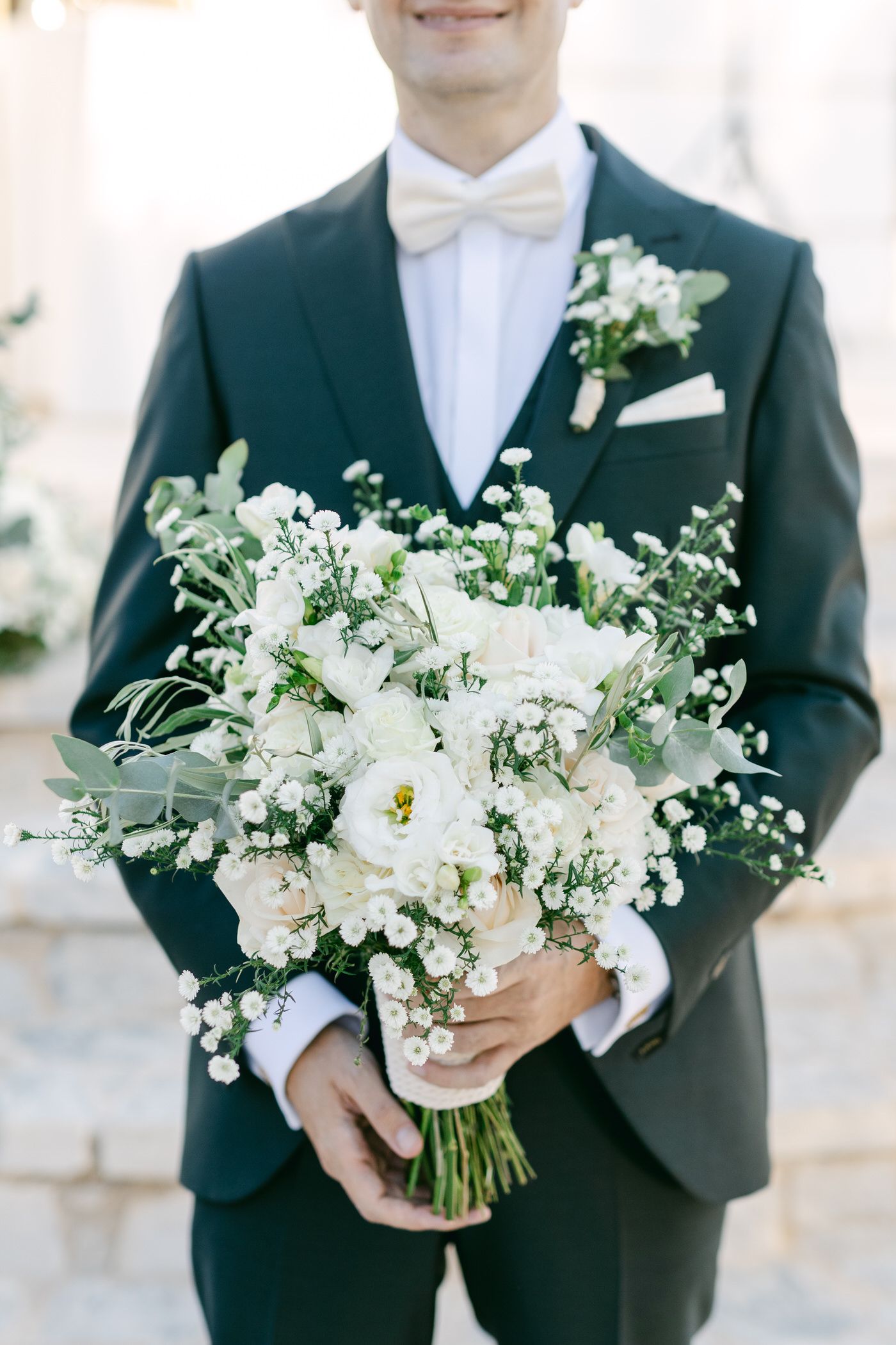 A stunning white wedding bouquet with delicate flowers and greenery held against a dark navy tuxedo and white bow tie.