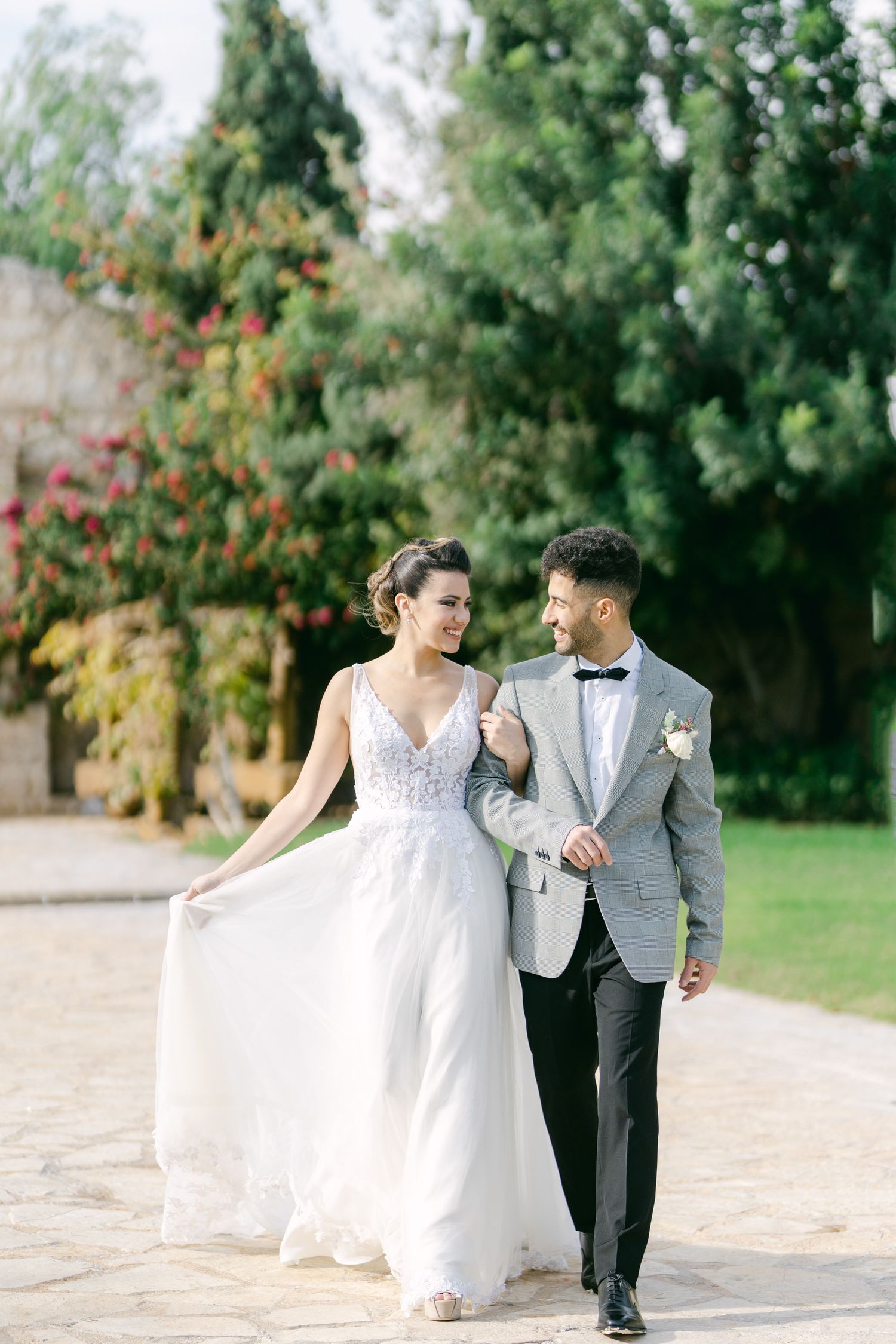 A wedding couple strolls along a garden path with the bride holding her flowing white dress.