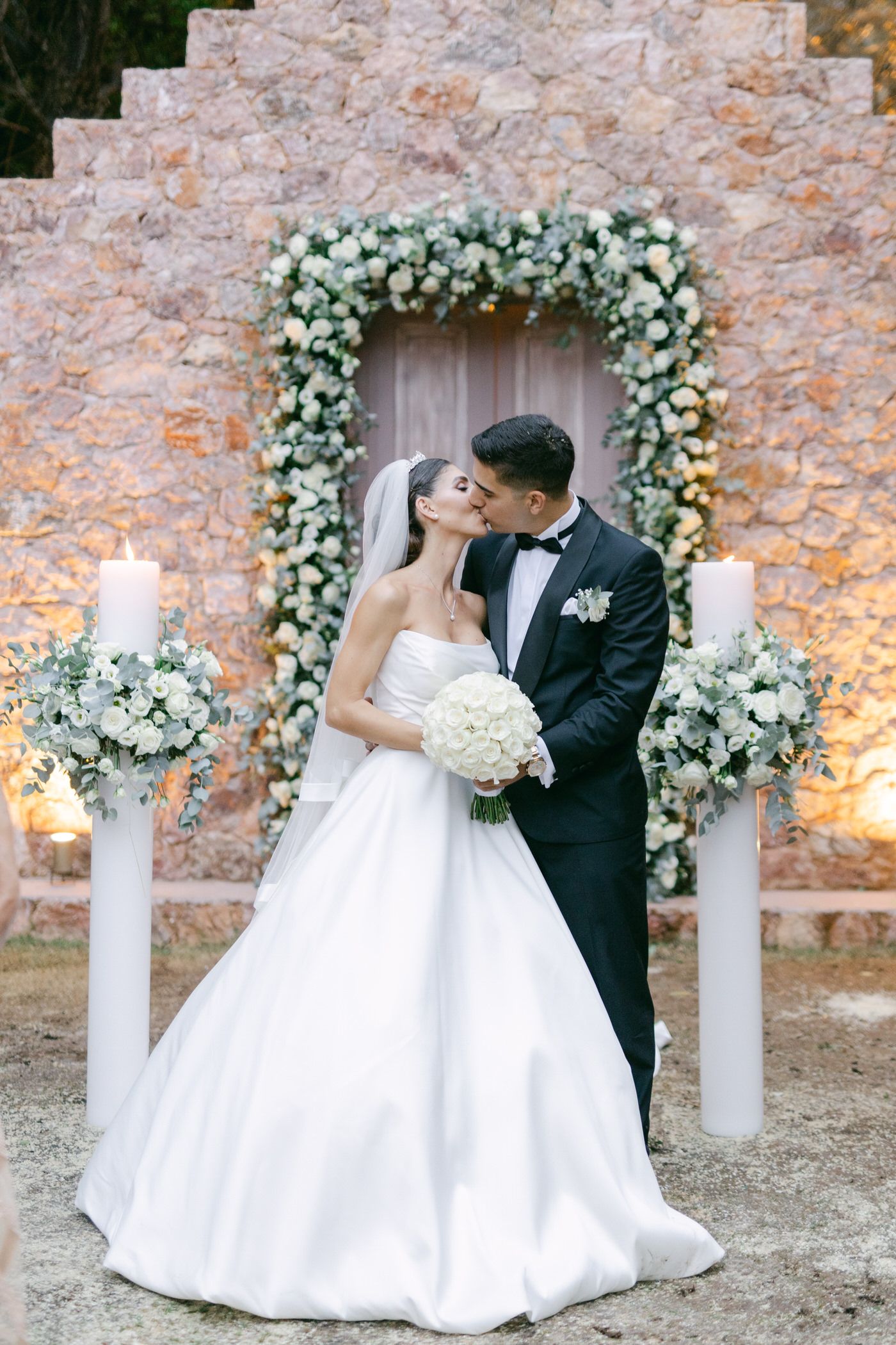 Elegant wedding scene with floral arch and white pillar candles against stone wall, featuring a couple in formal attire.