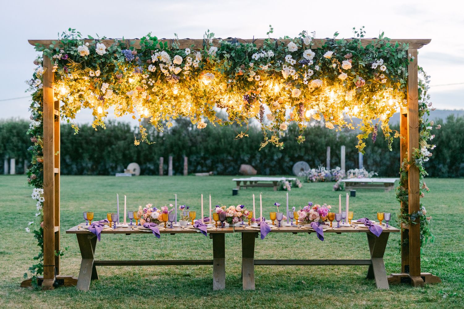 Rustic outdoor wedding reception table under wooden arch with twinkling lights and floral decorations in garden setting.