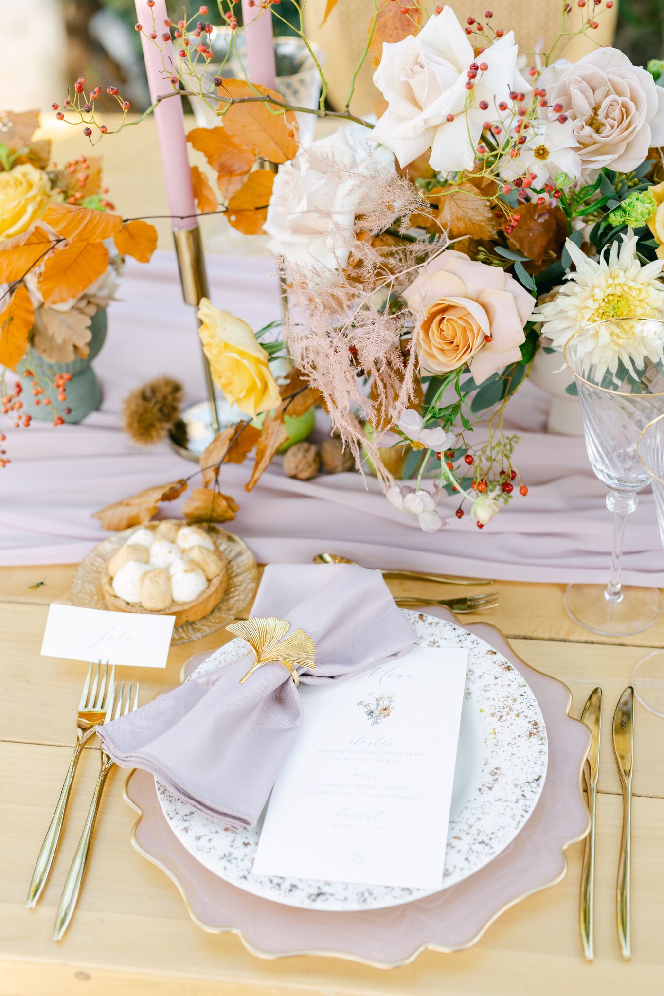 Elegant table setting with pastel roses, pumpkin-colored florals, and gold cutlery on lavender linens with scalloped plates.