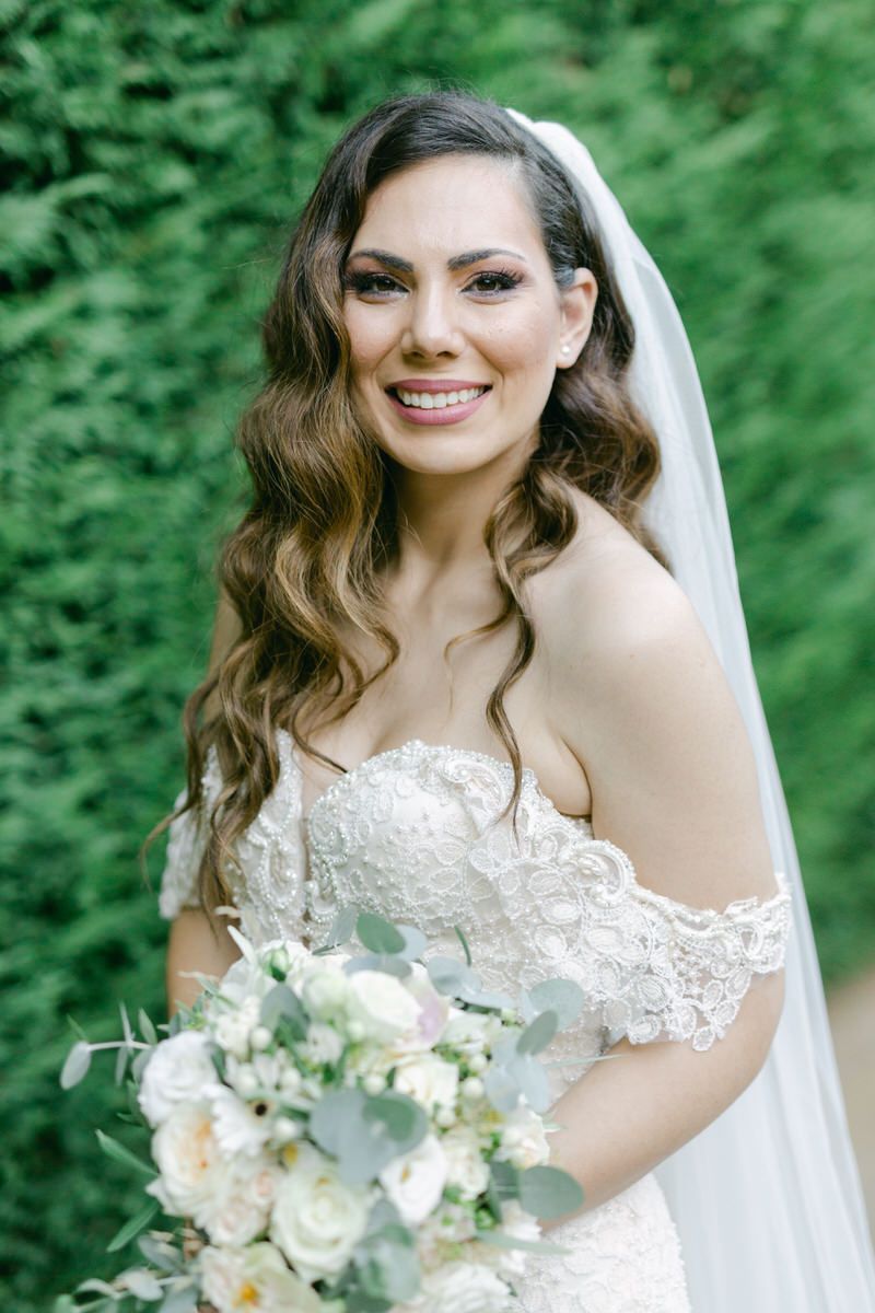 Beautiful bride in off-shoulder lace wedding dress holds pastel bouquet against green foliage backdrop.