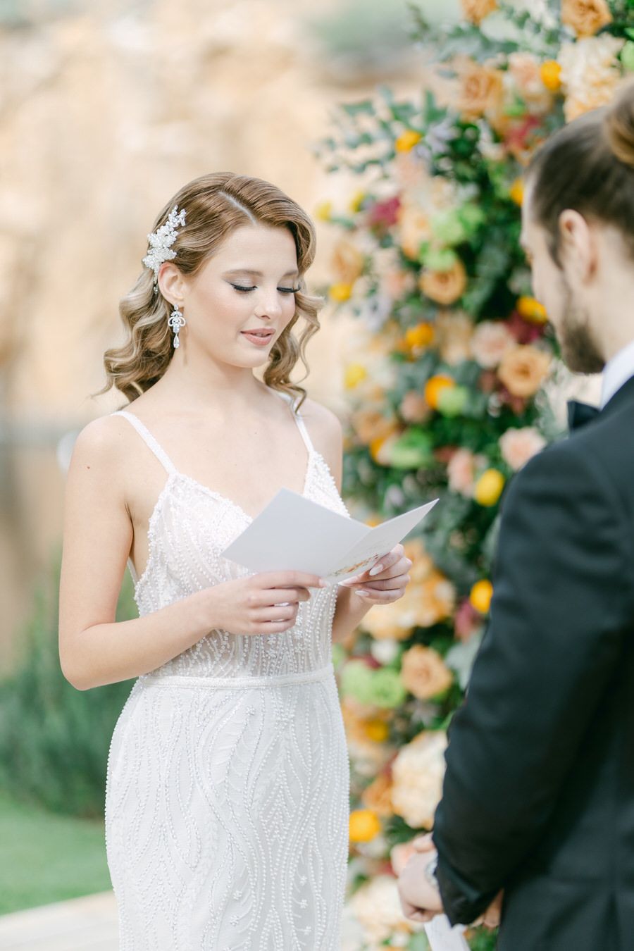 A bride in a lace gown reads vows in front of a floral arch decorated with peach roses.