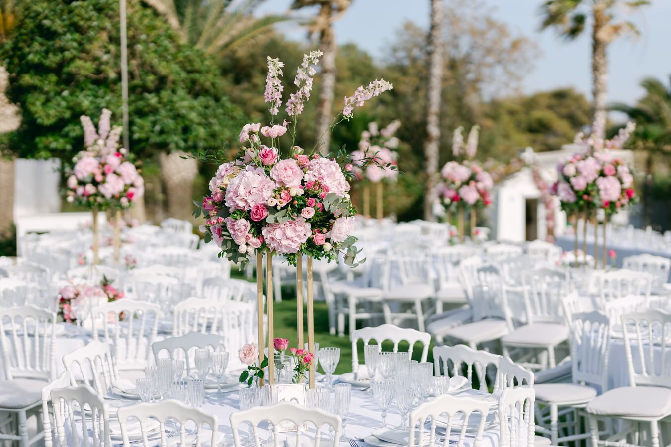 Elegant outdoor wedding reception setup with pink and white floral centerpieces on white tables.