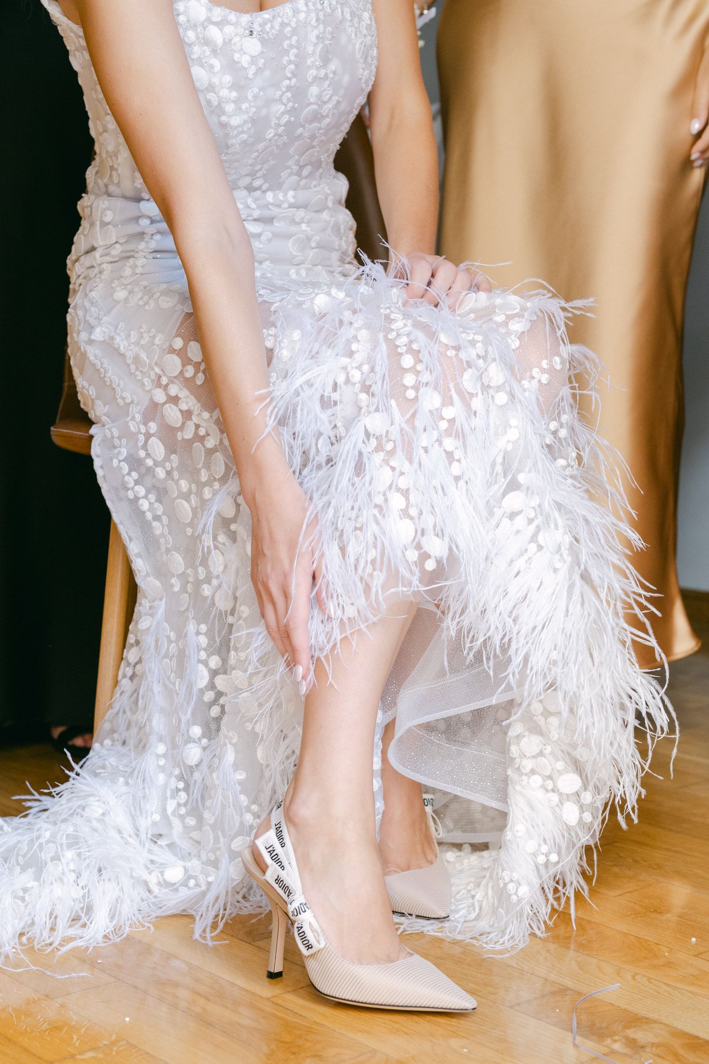 A detailed view of an elegant white feathered wedding gown and nude high heel shoes on a hardwood floor.