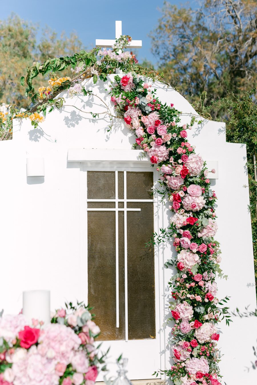 A white wedding arch decorated with pink roses and greenery against a natural outdoor backdrop features a cross on top.