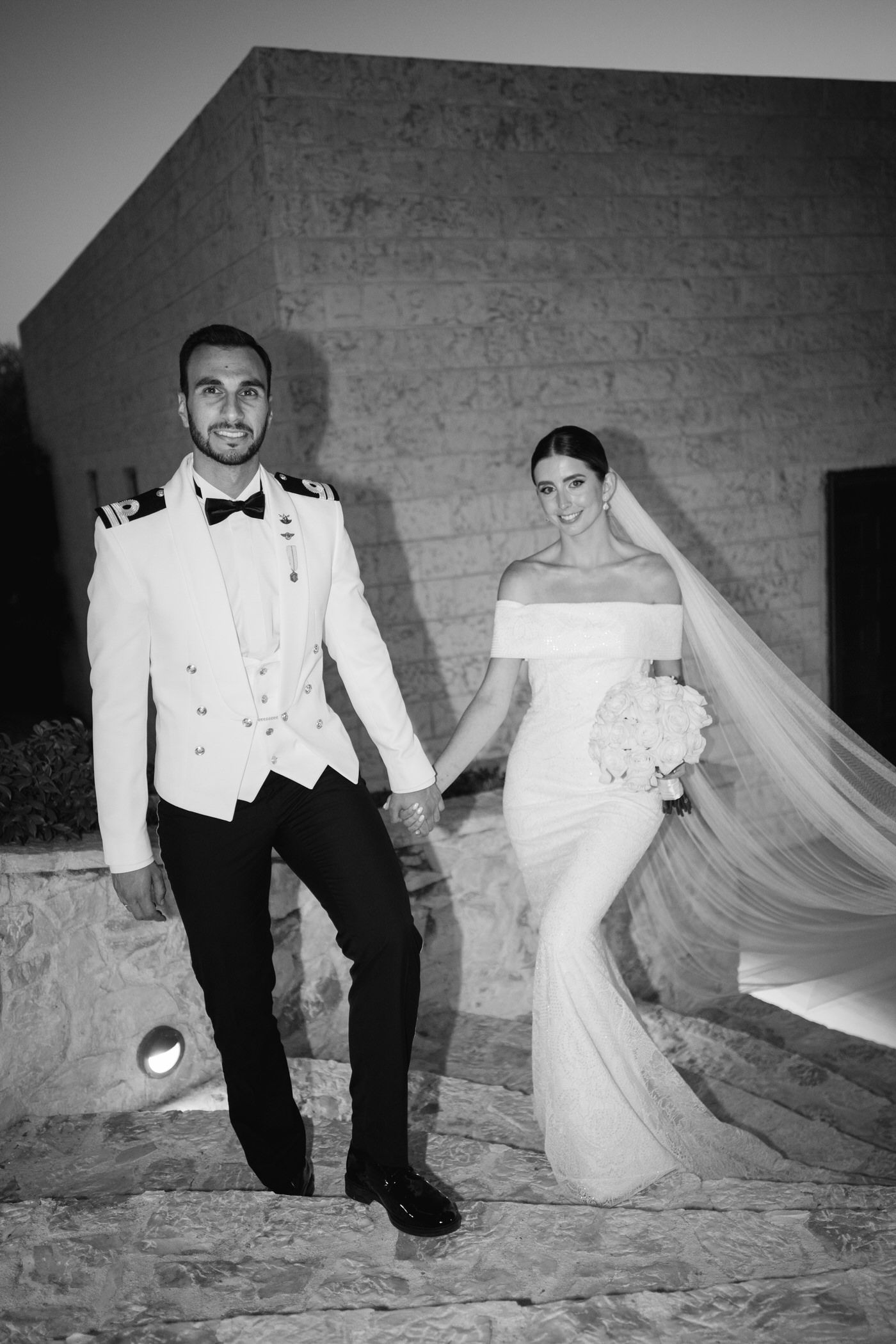 Black and white photo of a bride and groom holding hands outdoors, with bride wearing flowing veil and off-shoulder gown.