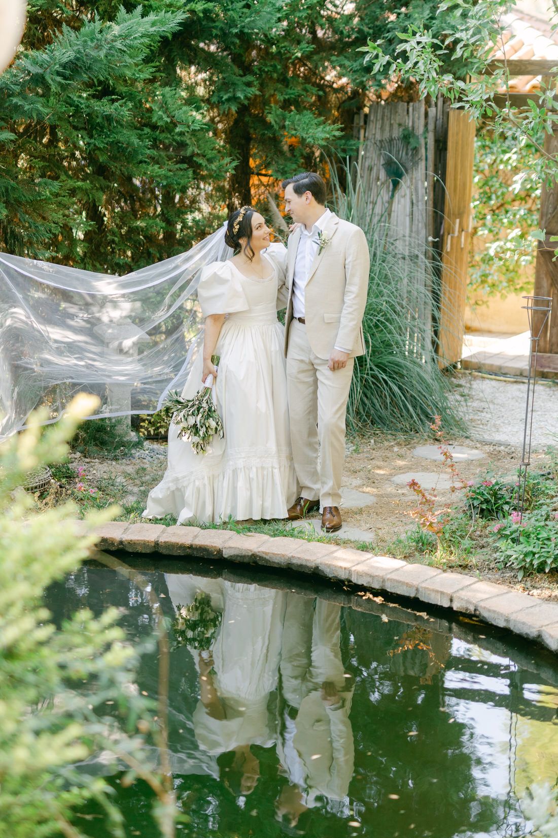 A romantic wedding couple stands on a curved bridge over a reflective pond in a lush garden setting with floating veil.