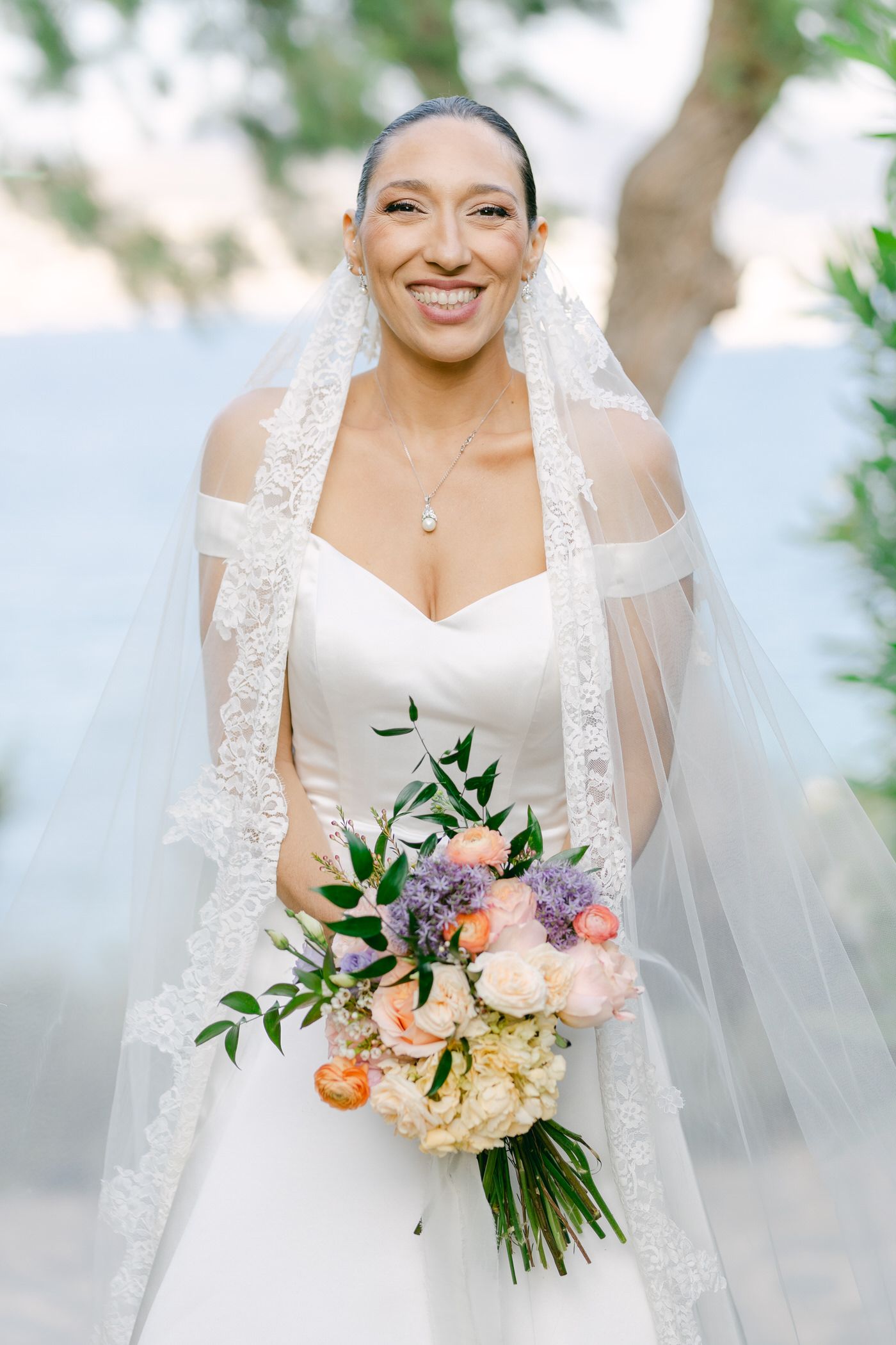 Beautiful bride in white wedding dress with lace veil holds colorful bouquet of roses and greenery on sunny day.
