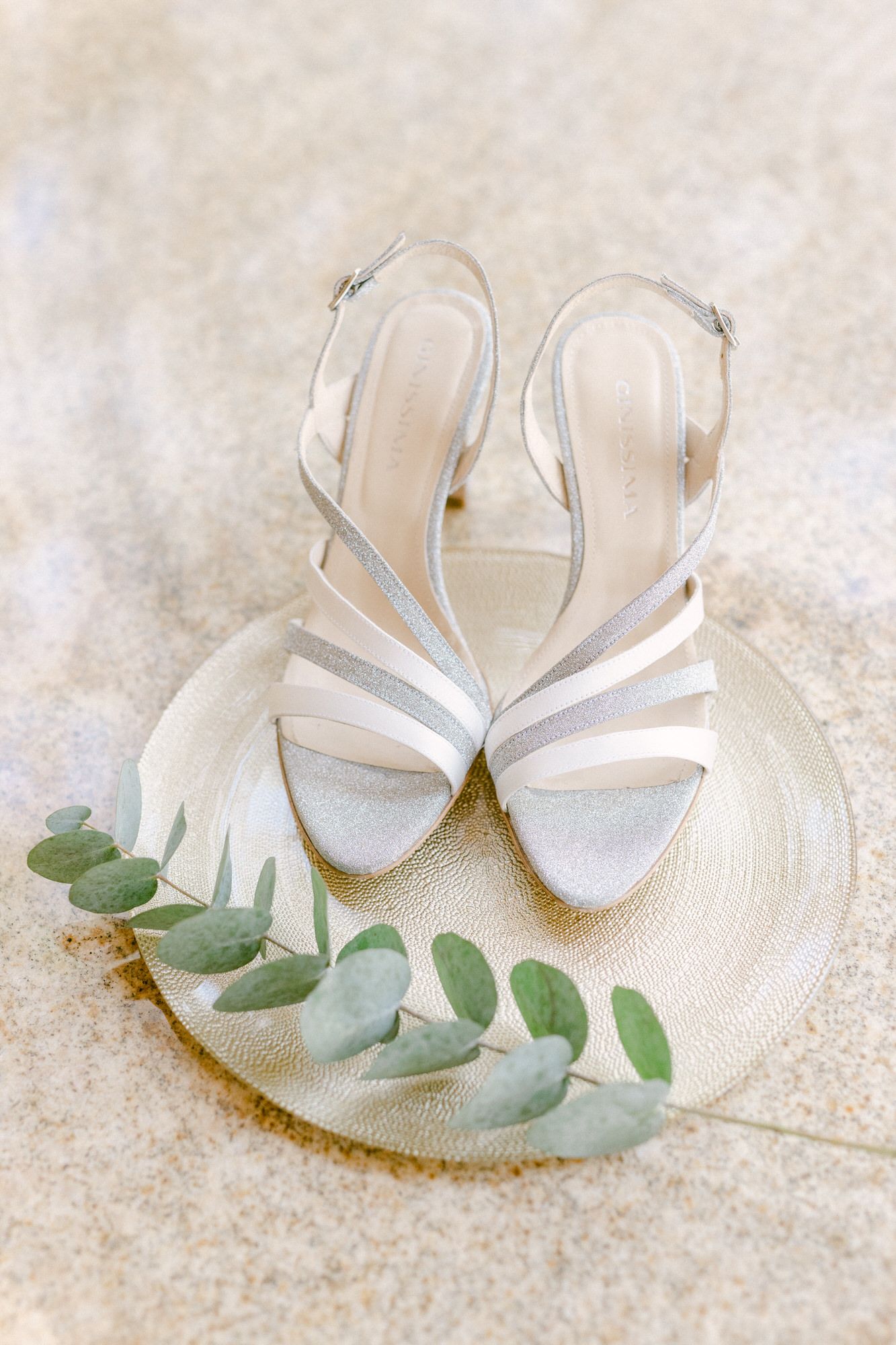 White strappy bridal sandals arranged with eucalyptus leaves on cream textured surface.