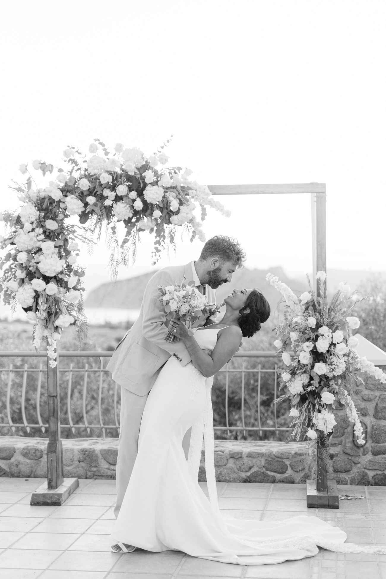 Couple sharing romantic moment under floral arch during outdoor wedding ceremony with mountain backdrop.