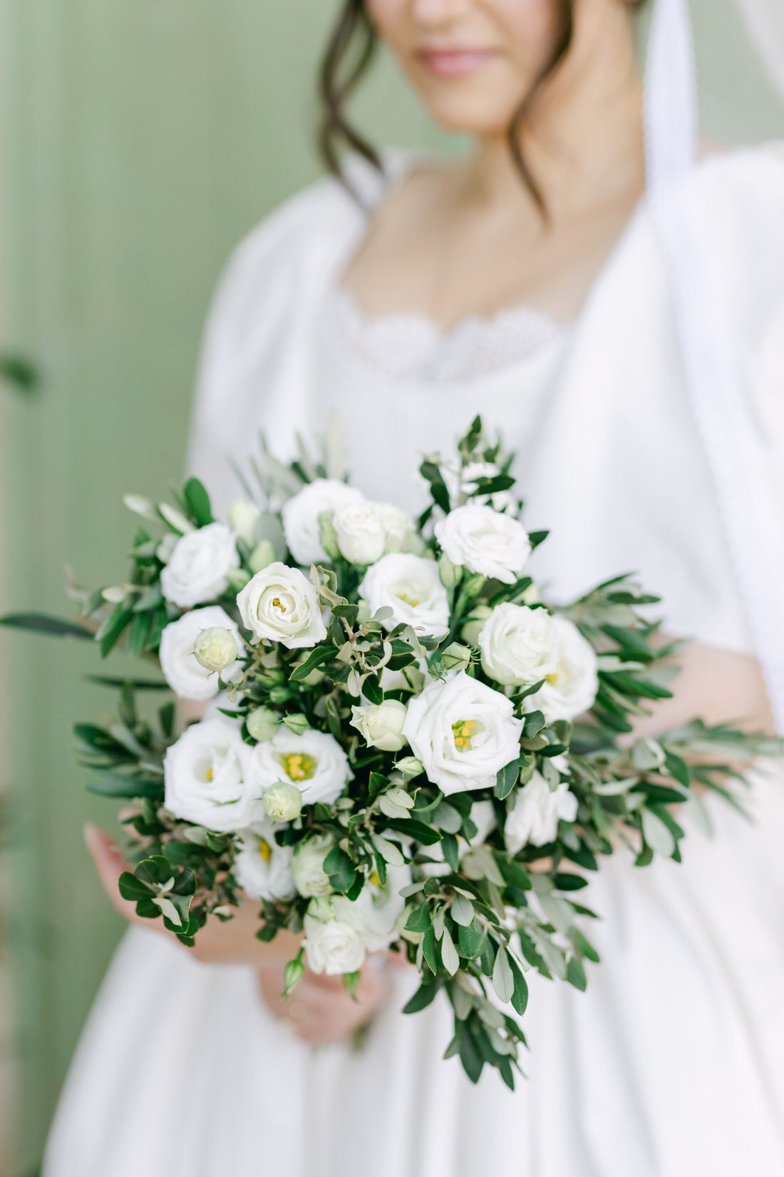 Beautiful white rose and greenery wedding bouquet held against a flowing white dress with soft green background.