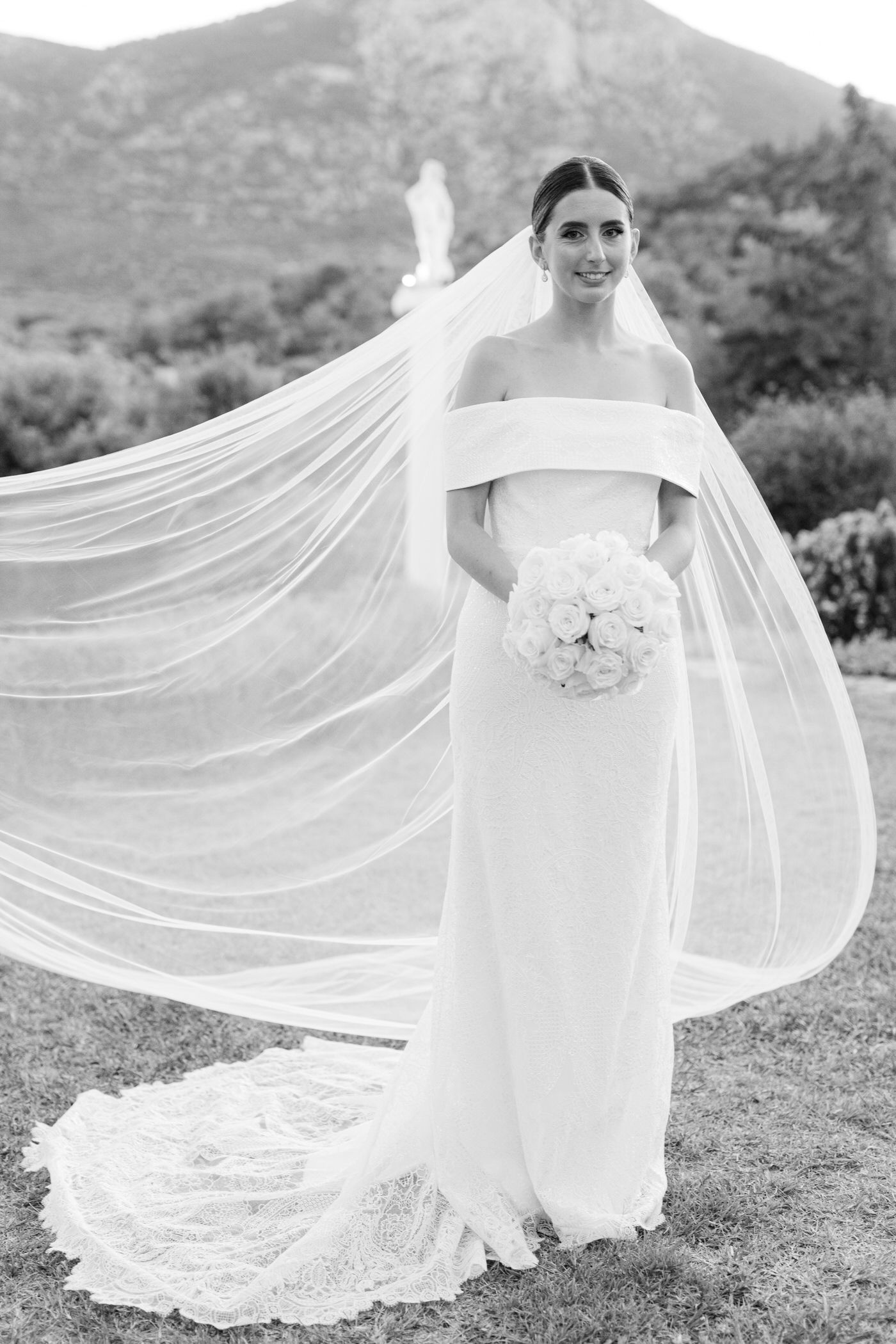 Elegant black and white wedding photo featuring bride in off-shoulder gown with flowing veil against mountain backdrop.