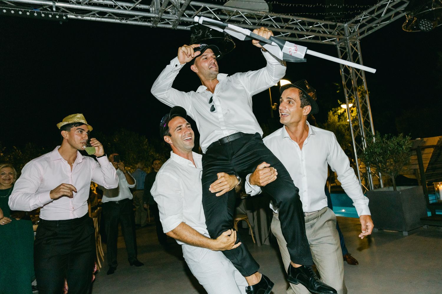 Group of party guests dance energetically at a nighttime reception, with festive lighting and staging visible in background. Γαμήλιο πάρτυ.