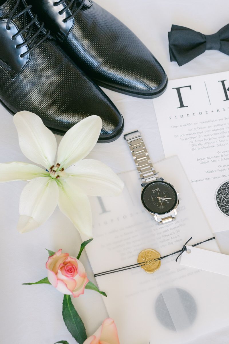 Elegant black leather dress shoes arranged with a bow tie, watch, white lily and pink rose on white paper.