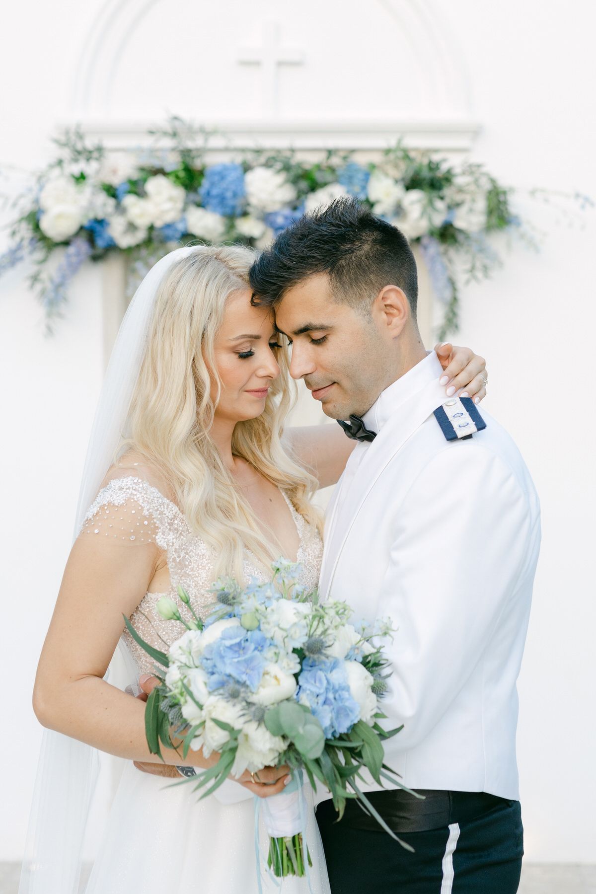 A romantic wedding couple holds a blue and white floral bouquet against a flower-adorned backdrop.