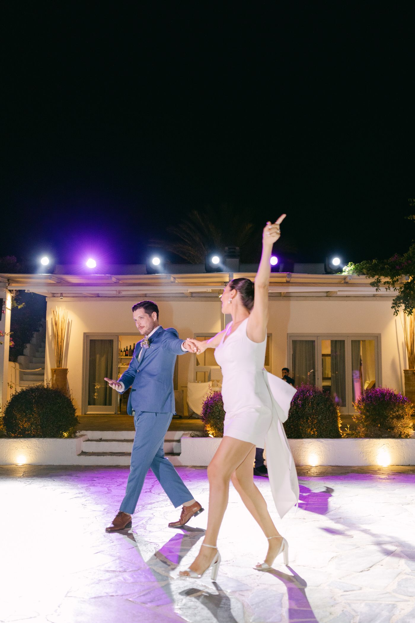 A couple dances energetically at an outdoor venue illuminated by purple and white lights during their wedding reception.