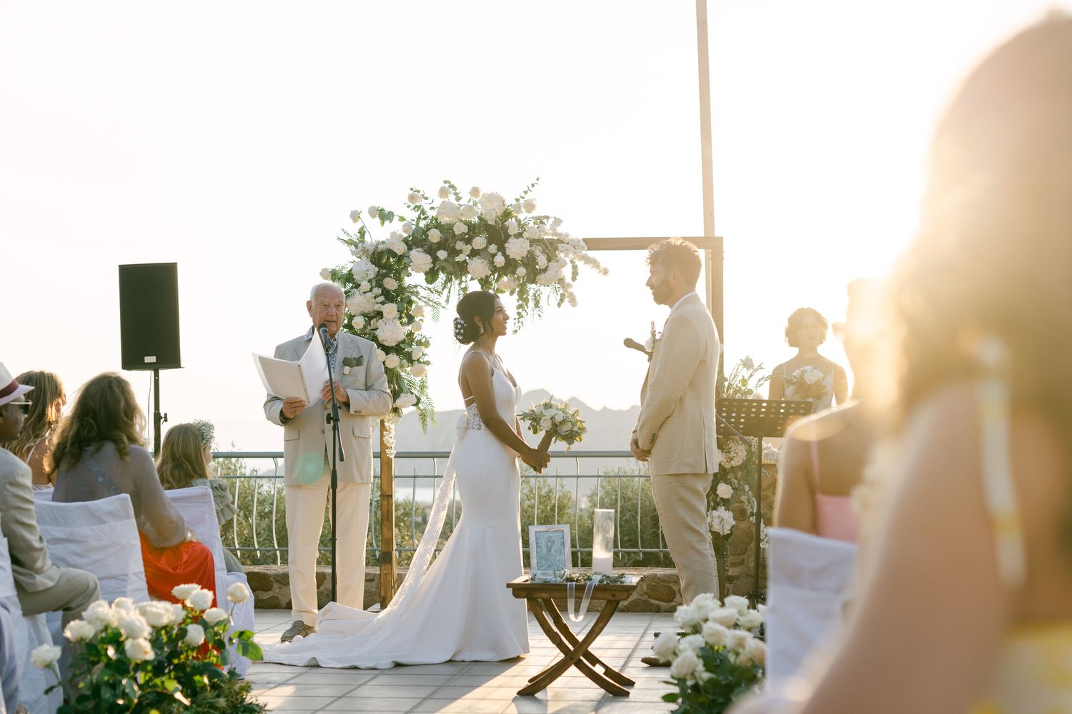 Outdoor wedding ceremony on terrace with floral arch and guests at sunset.
