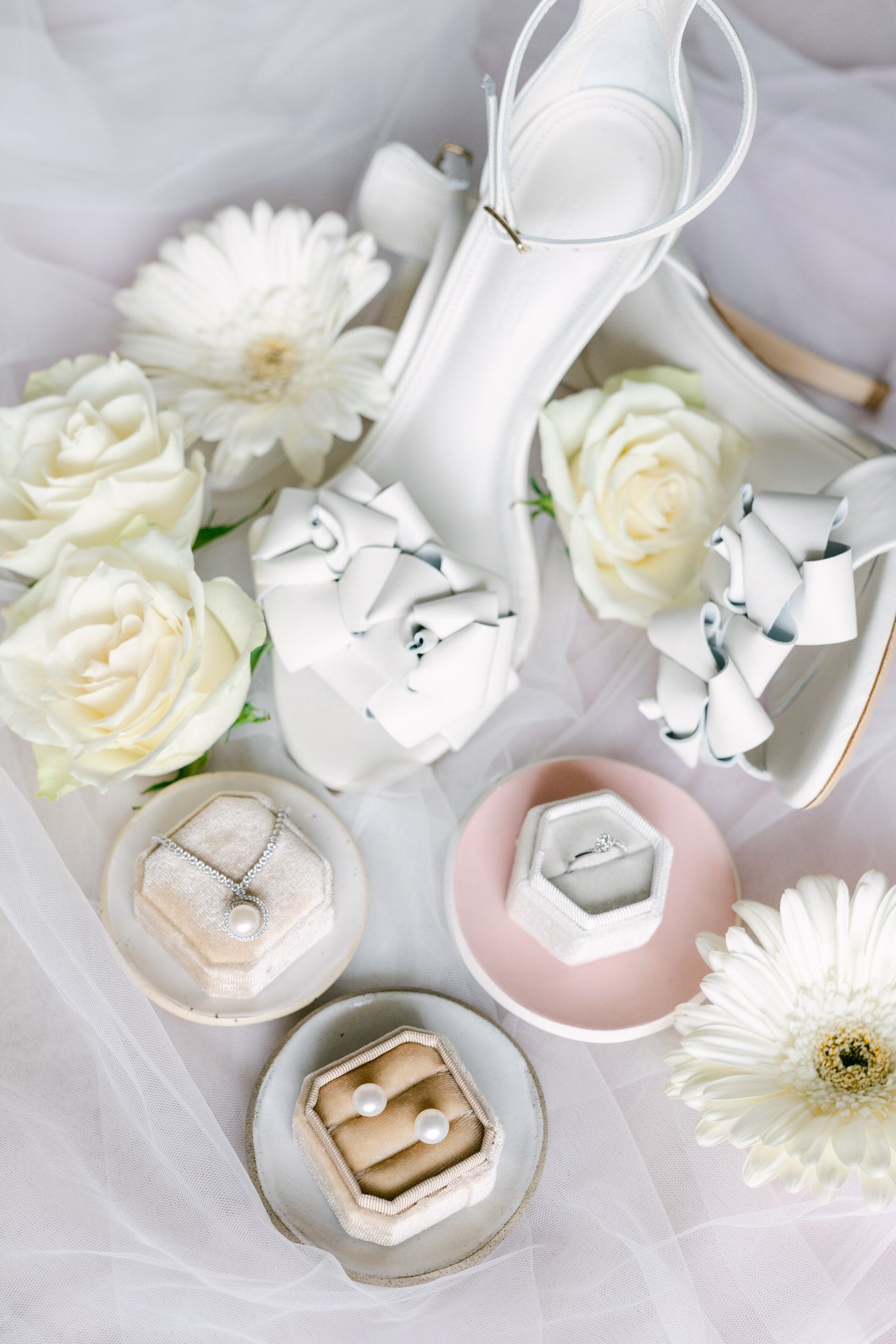 A bridal themed flatlay with white roses, ring boxes in pastel colors, and a white heel shoe on soft fabric.