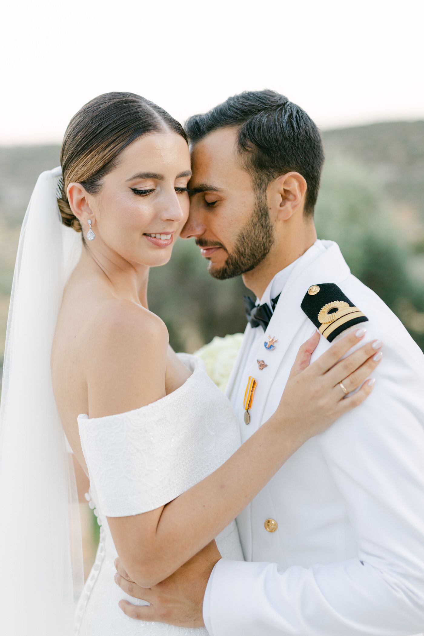 A bride in white gown and groom in naval uniform share an intimate moment during their wedding photoshoot outdoors.