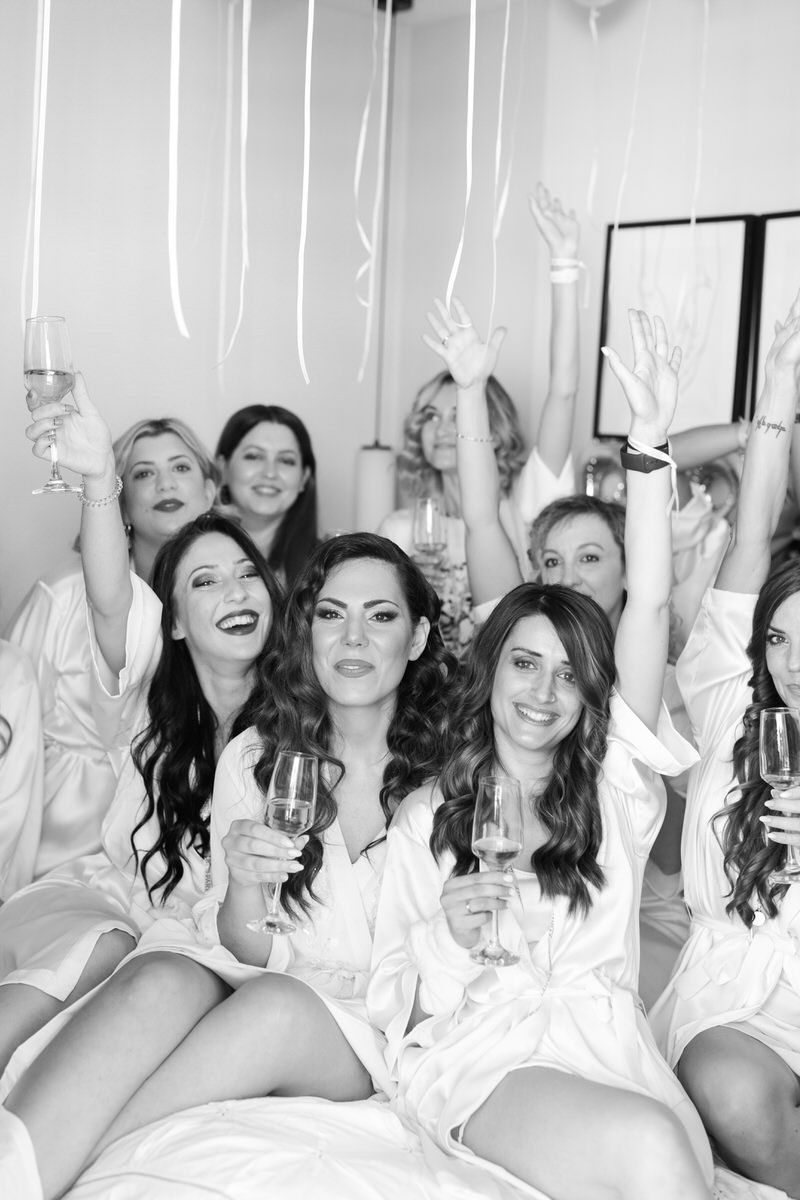 A group in white silk robes raise their champagne glasses in celebration while sitting on a bed together.