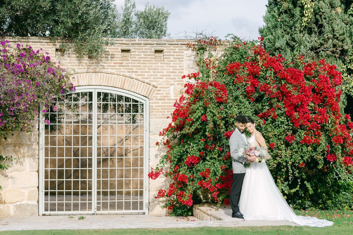 Wedding couple posing by a stone gate with cascading red roses.