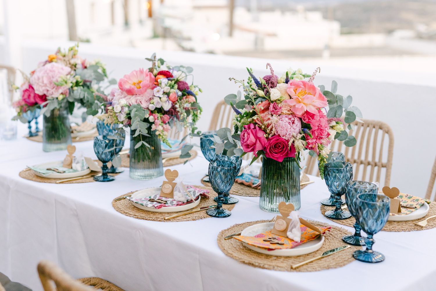 Elegant wedding table setting with pink floral centerpieces, blue goblets, and rattan chargers on white linen tablecloth.