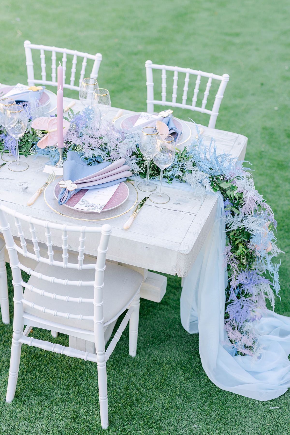 Elegant outdoor table setting with white Chiavari chairs, floral garland, and soft blue accents on a grassy lawn.
