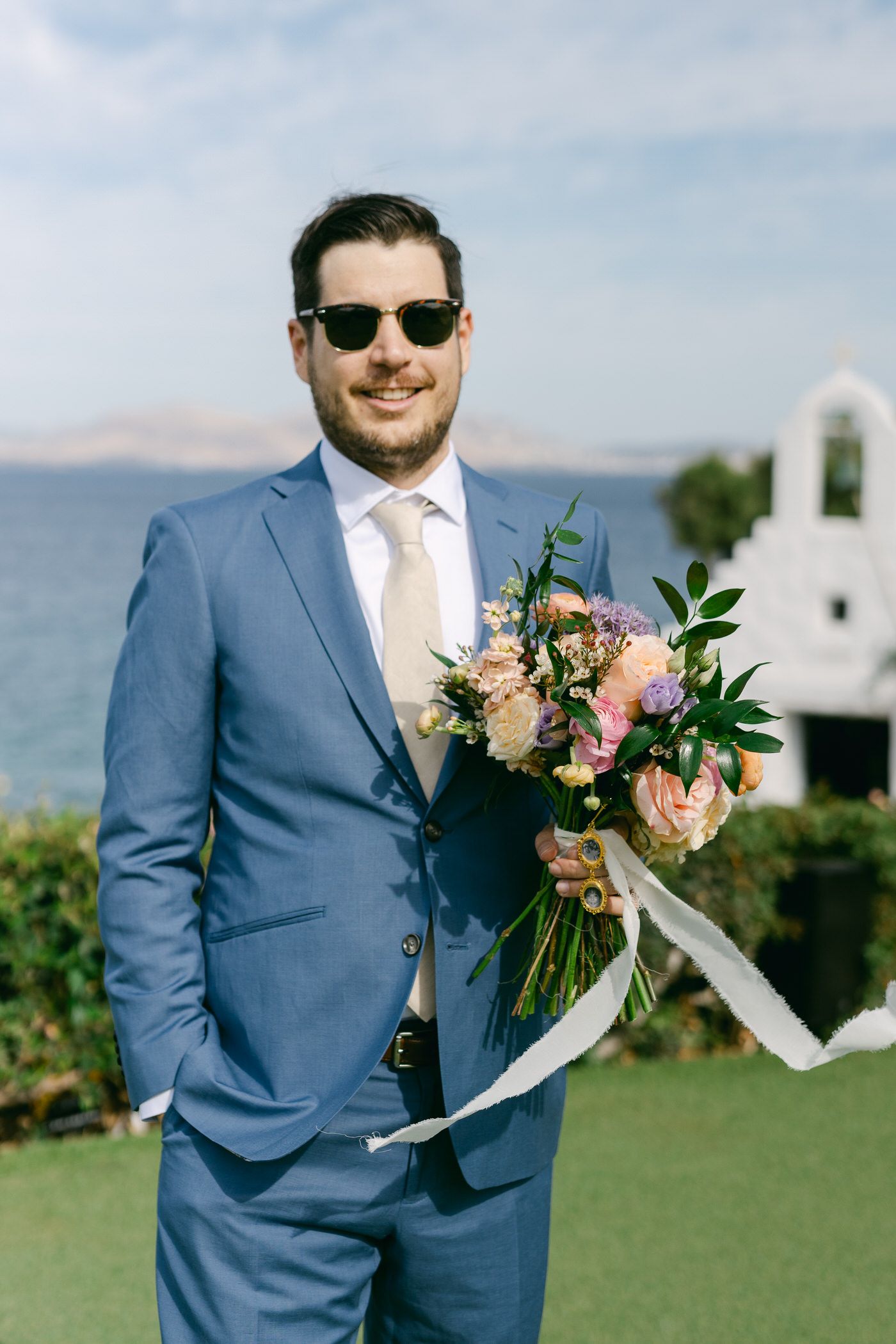 A groom in a light blue suit and sunglasses holds a blush and peach floral bouquet against a coastal backdrop.