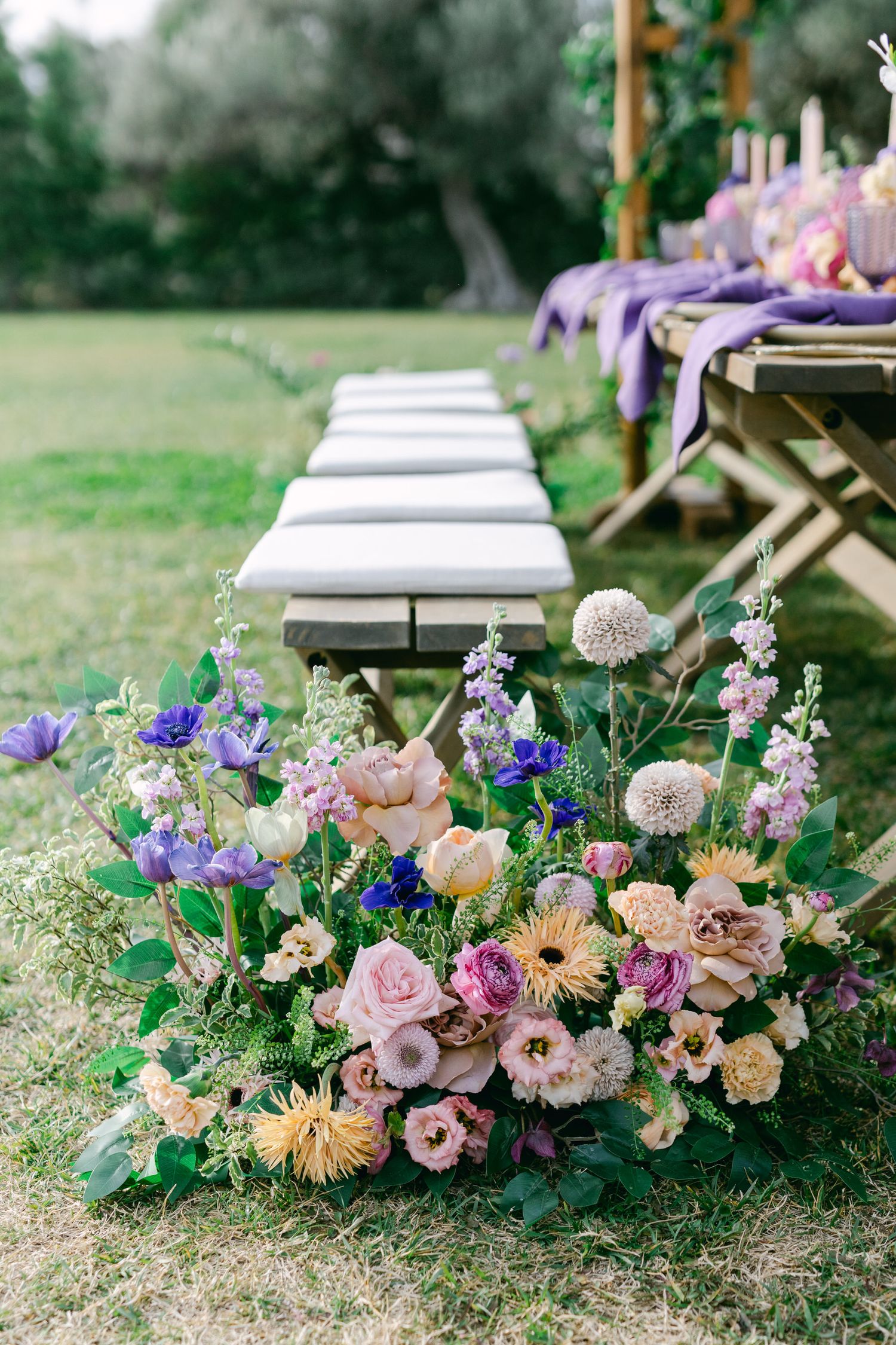 Beautiful floral arrangement with pink, purple and peach blooms decorates an outdoor event setup with wooden benches.