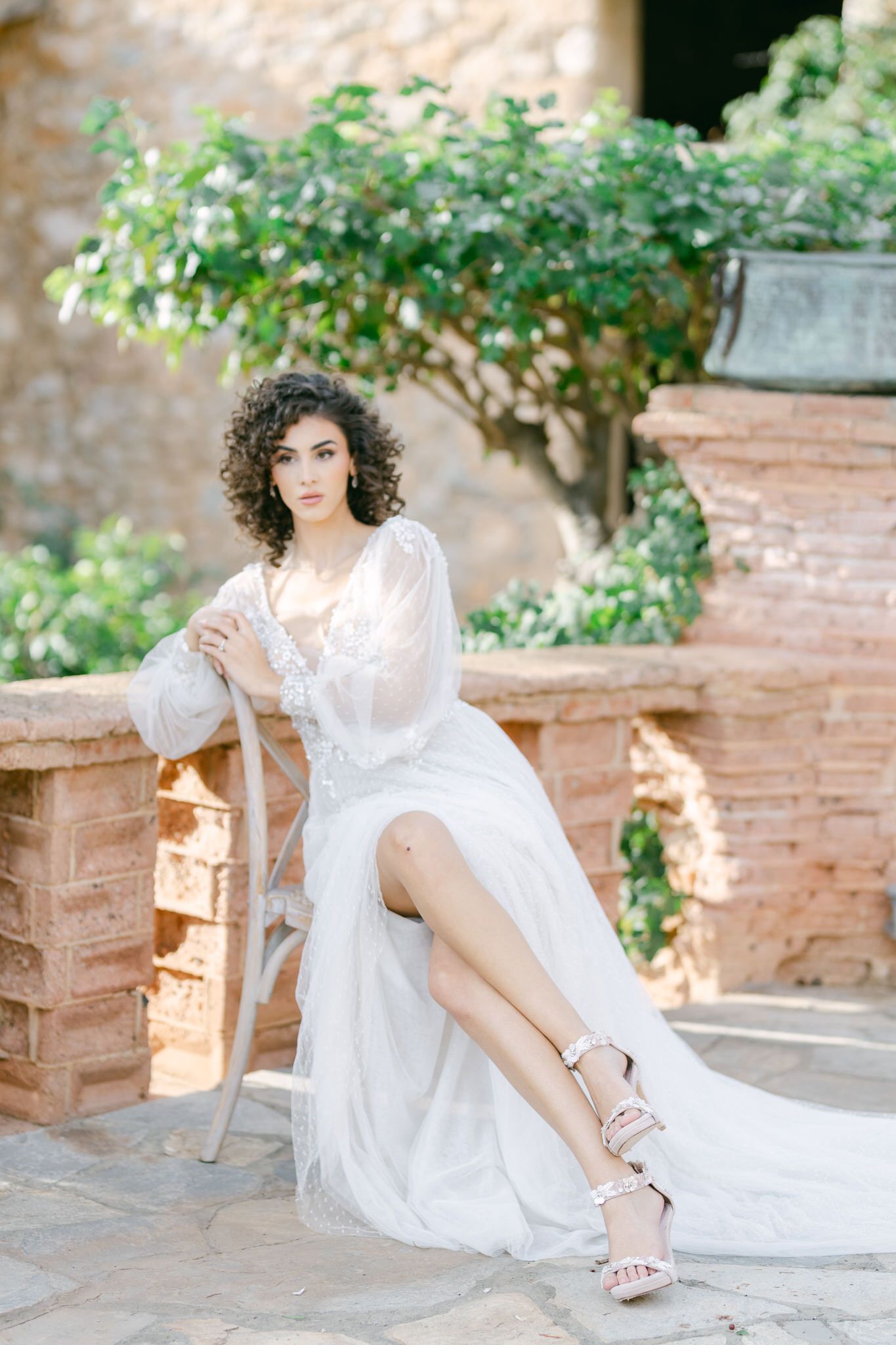 Elegant white flowy dress with romantic styling photographed in a Mediterranean garden setting with brick walls and greenery.