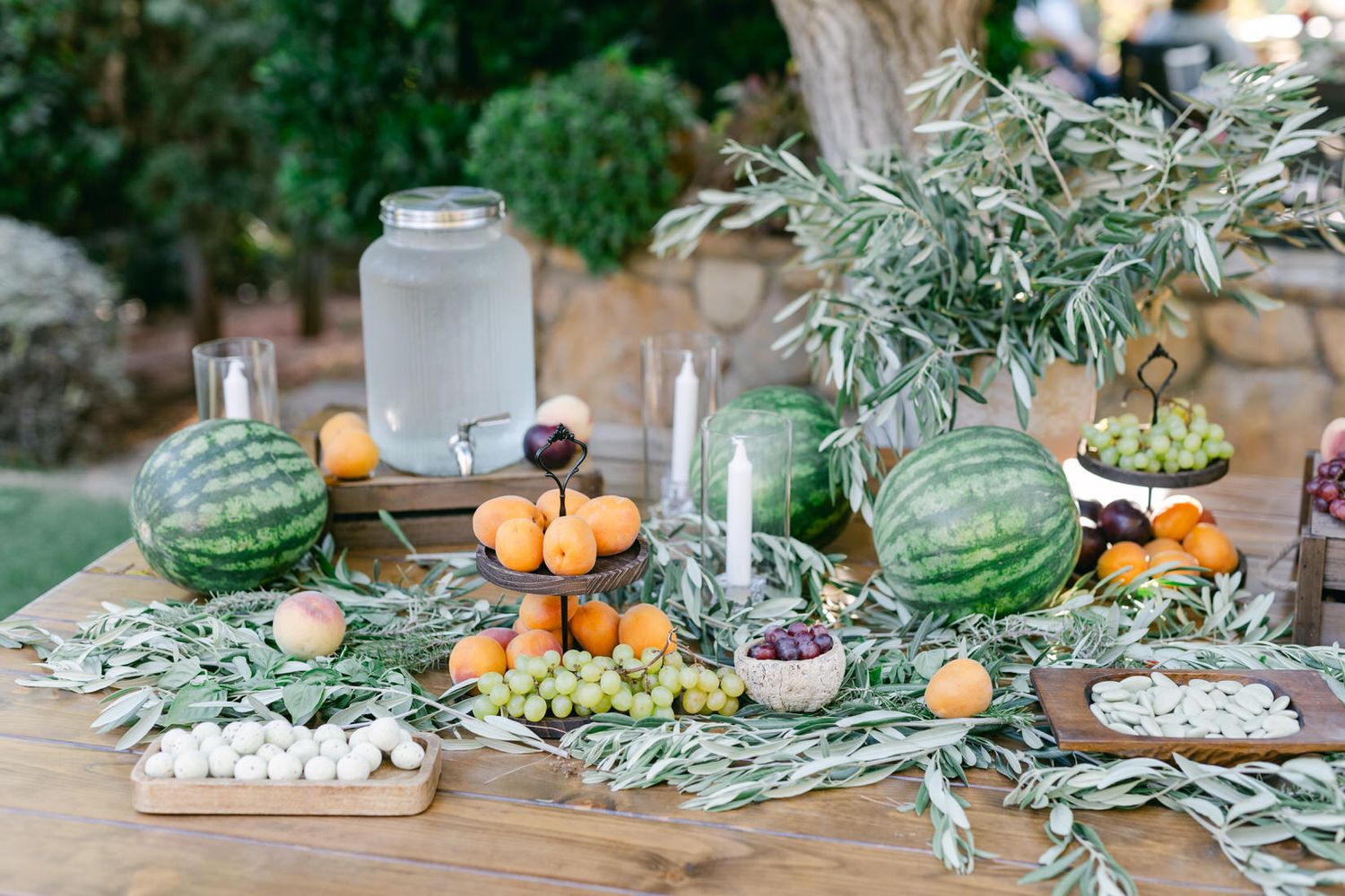 Summer outdoor table setting with watermelons, citrus fruits, candles and olive branches on rustic wooden table.