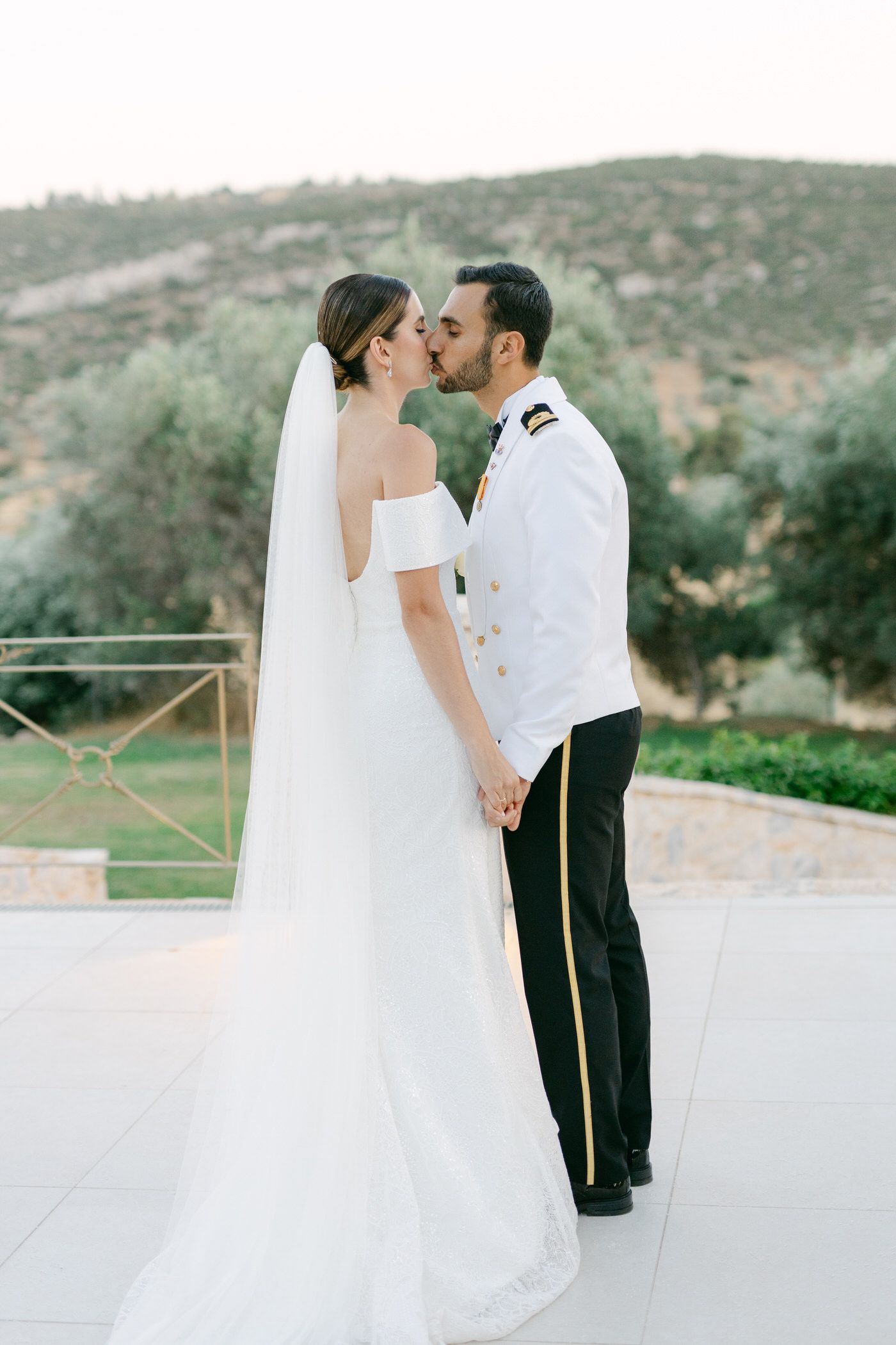 A romantic wedding couple shares a kiss on an outdoor terrace with rolling hills in the background at dusk.