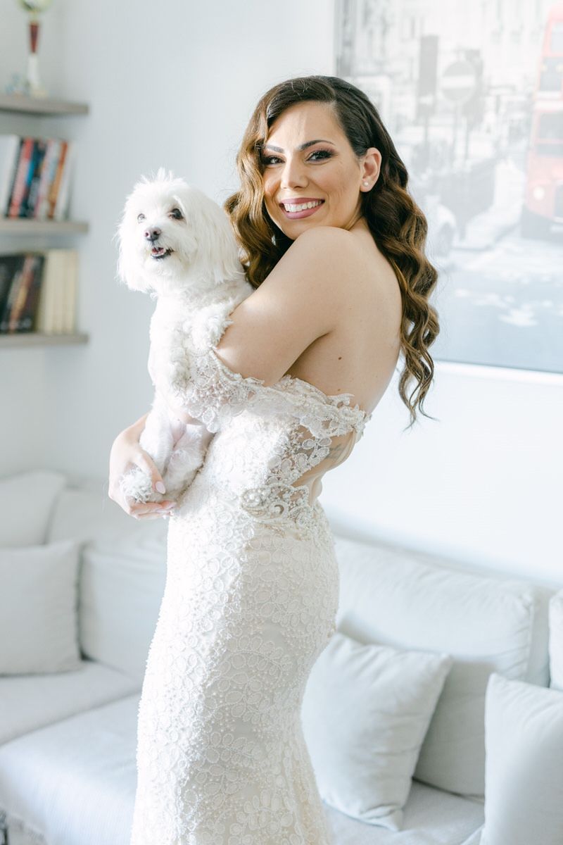 A bride in a fitted lace gown holds a small white fluffy dog while standing in a bright white room.