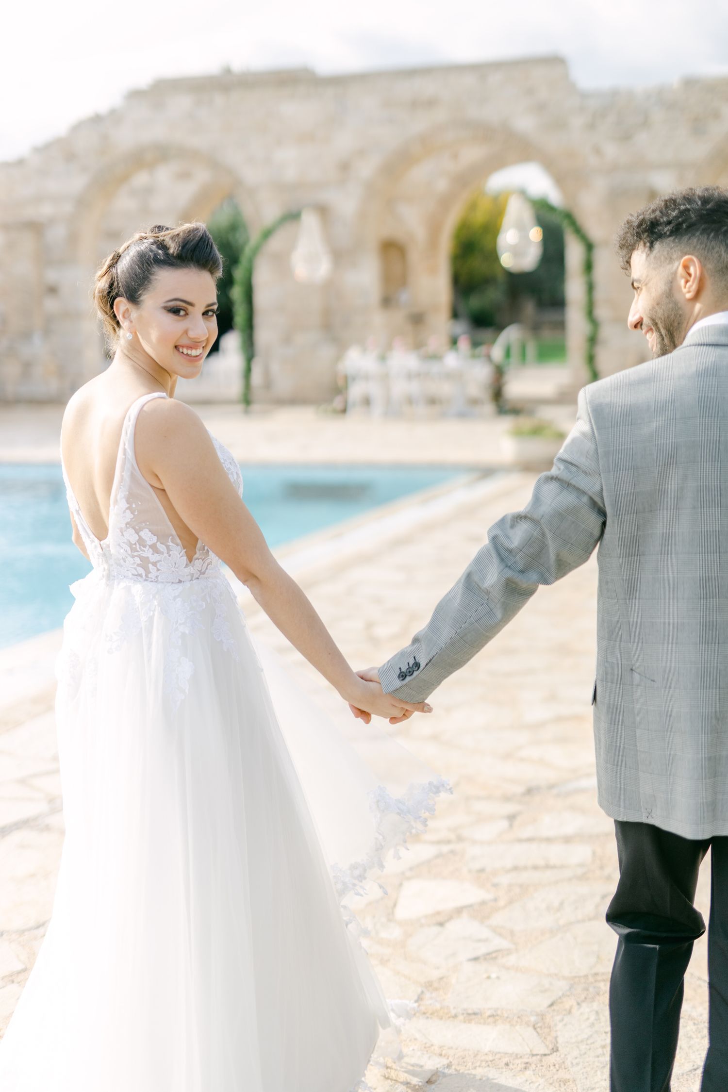 Wedding couple holding hands by a pool at a stone venue with romantic architecture.