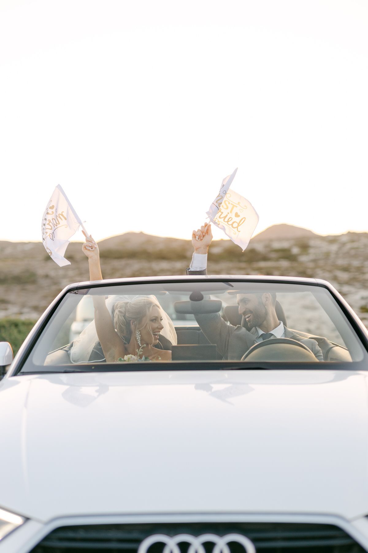 A white Audi convertible decorated with wedding flags drives newly married couple through scenic landscape.