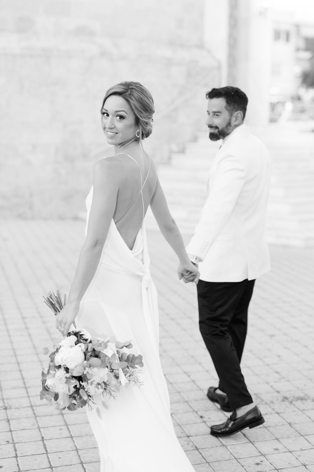 A bride in an elegant backless white gown holds hands with her groom in a white jacket during a black and white photoshoot.