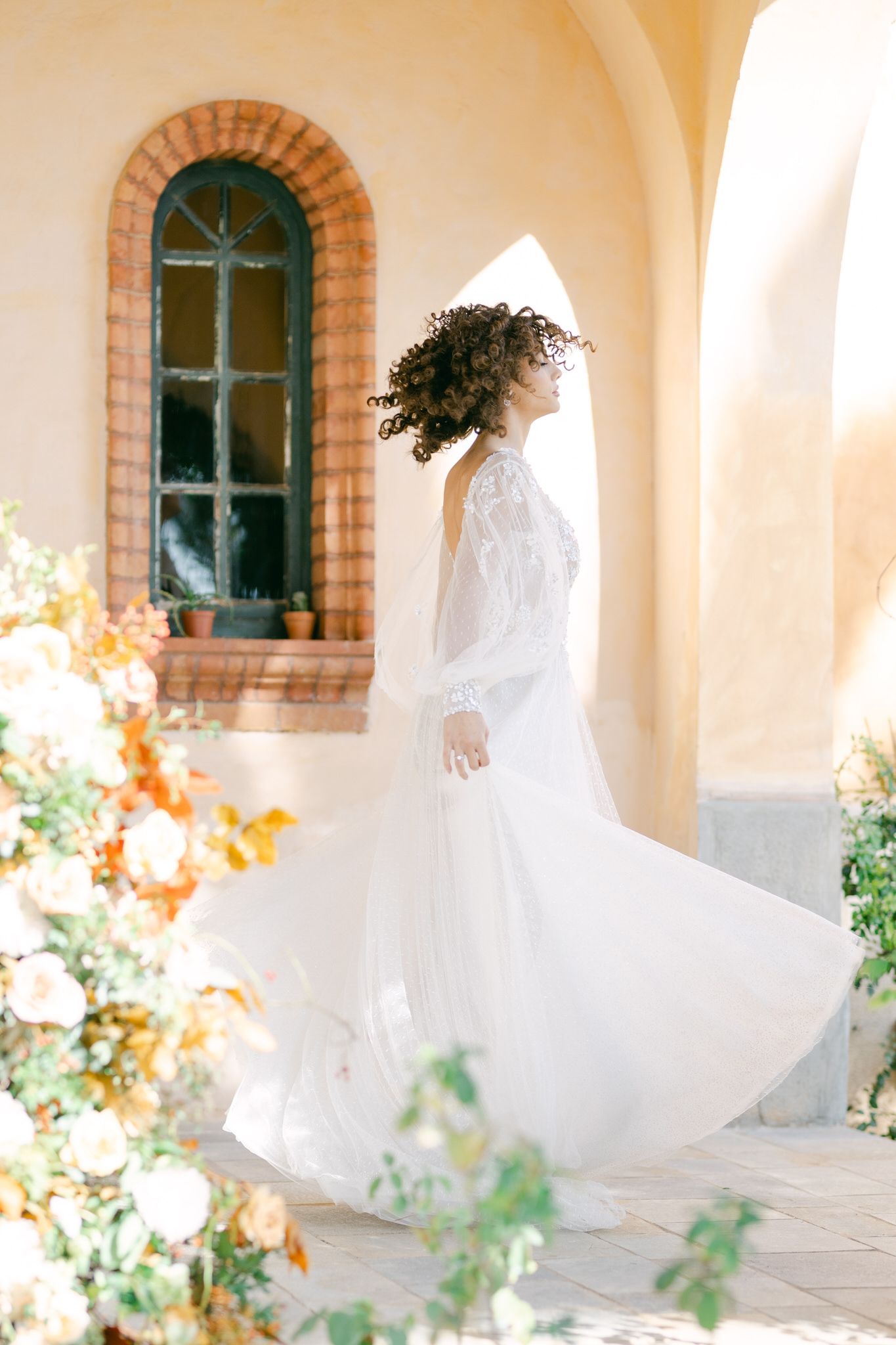 Ethereal white wedding dress swirling in motion against an arched window and floral backdrop.