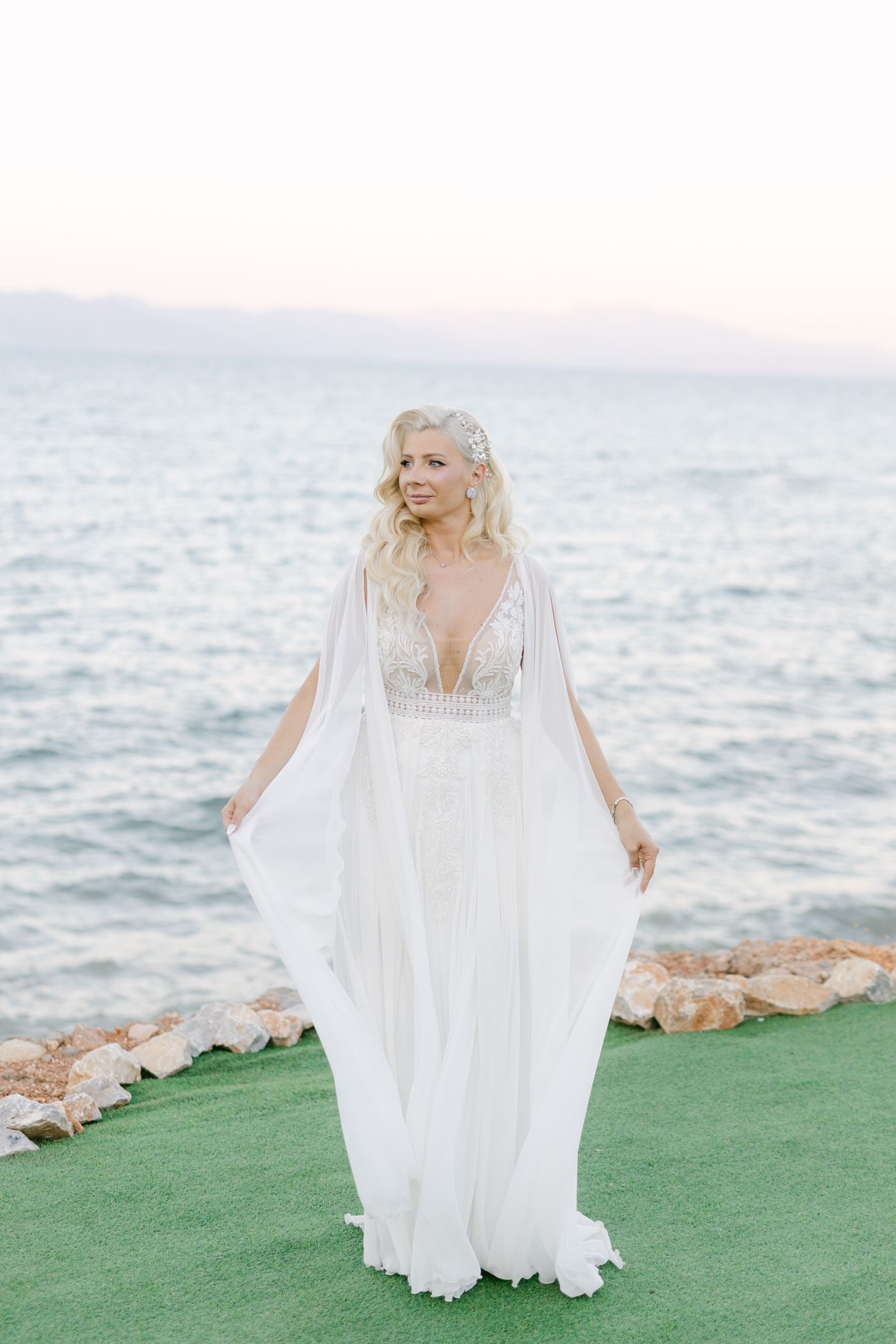 Flowing white wedding dress with cape sleeves photographed against ocean backdrop on green lawn.