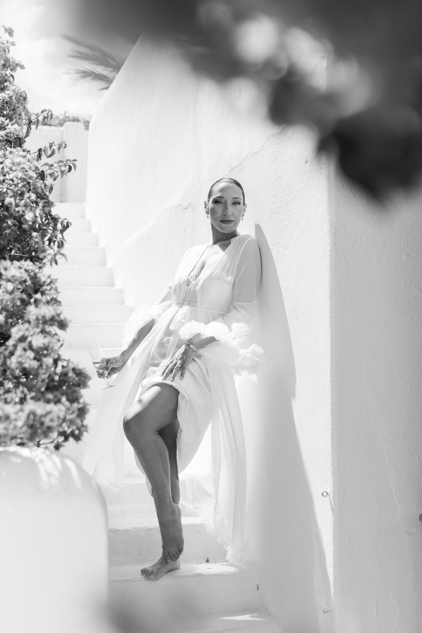 Artistic black and white fashion photograph with soft lighting showing a figure in flowing white clothing against a wall.
